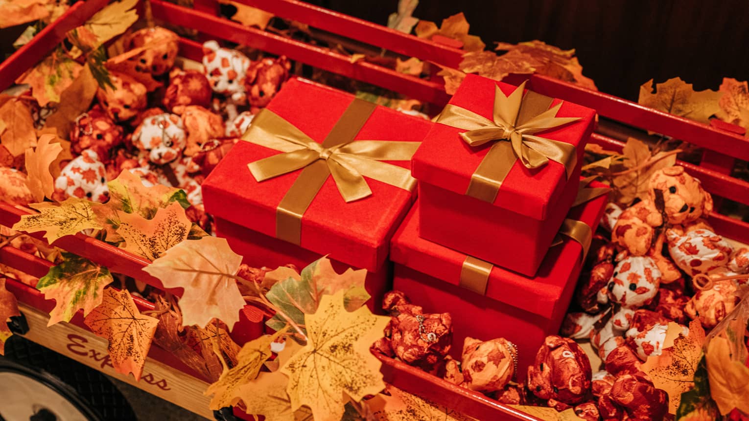 Three red gift boxes wrapped in gold ribbons sit in a wagon filled with orange leaves