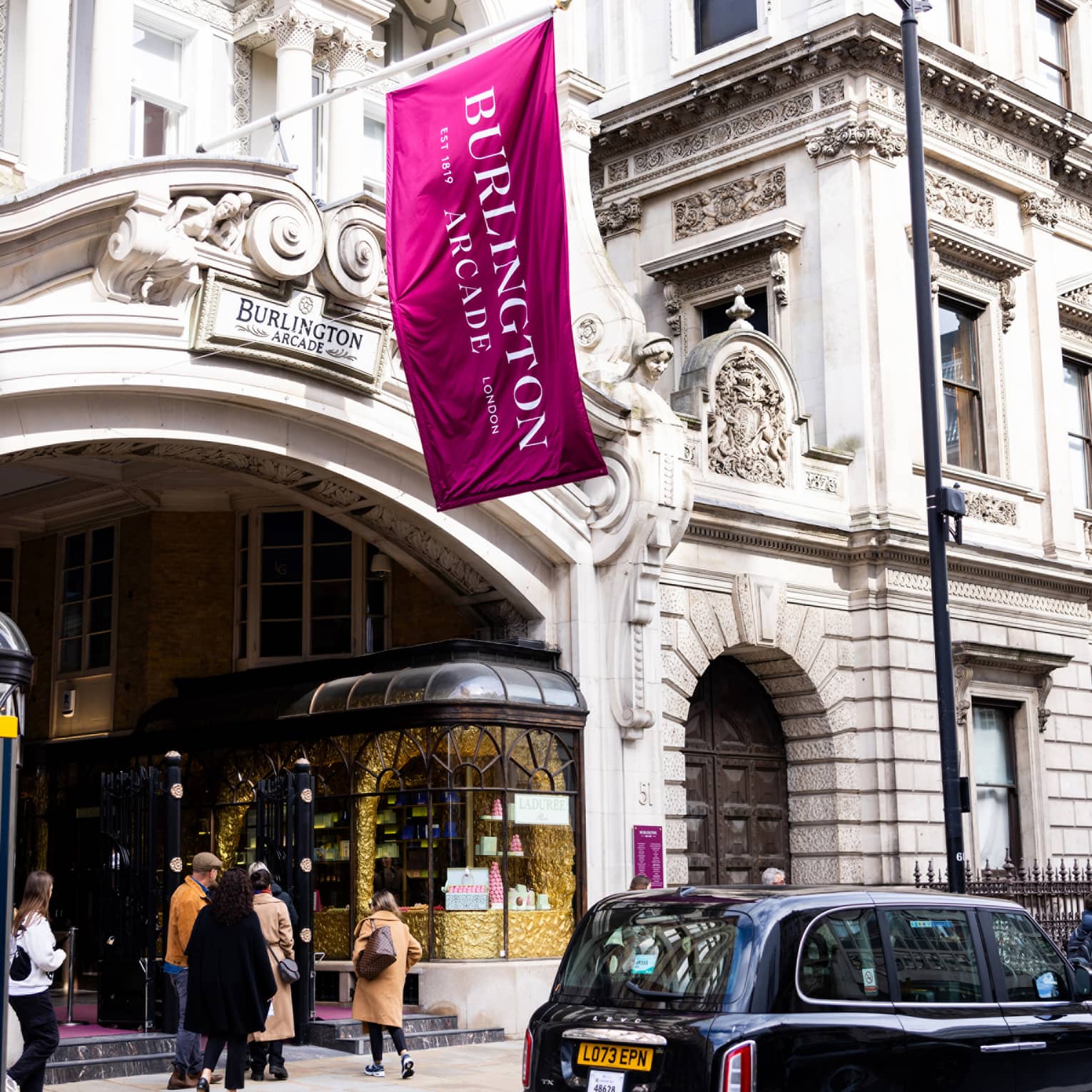 London-style black cab parked in front of a high-end pastry shop in a building with a Burlington Arcade sign and flag.