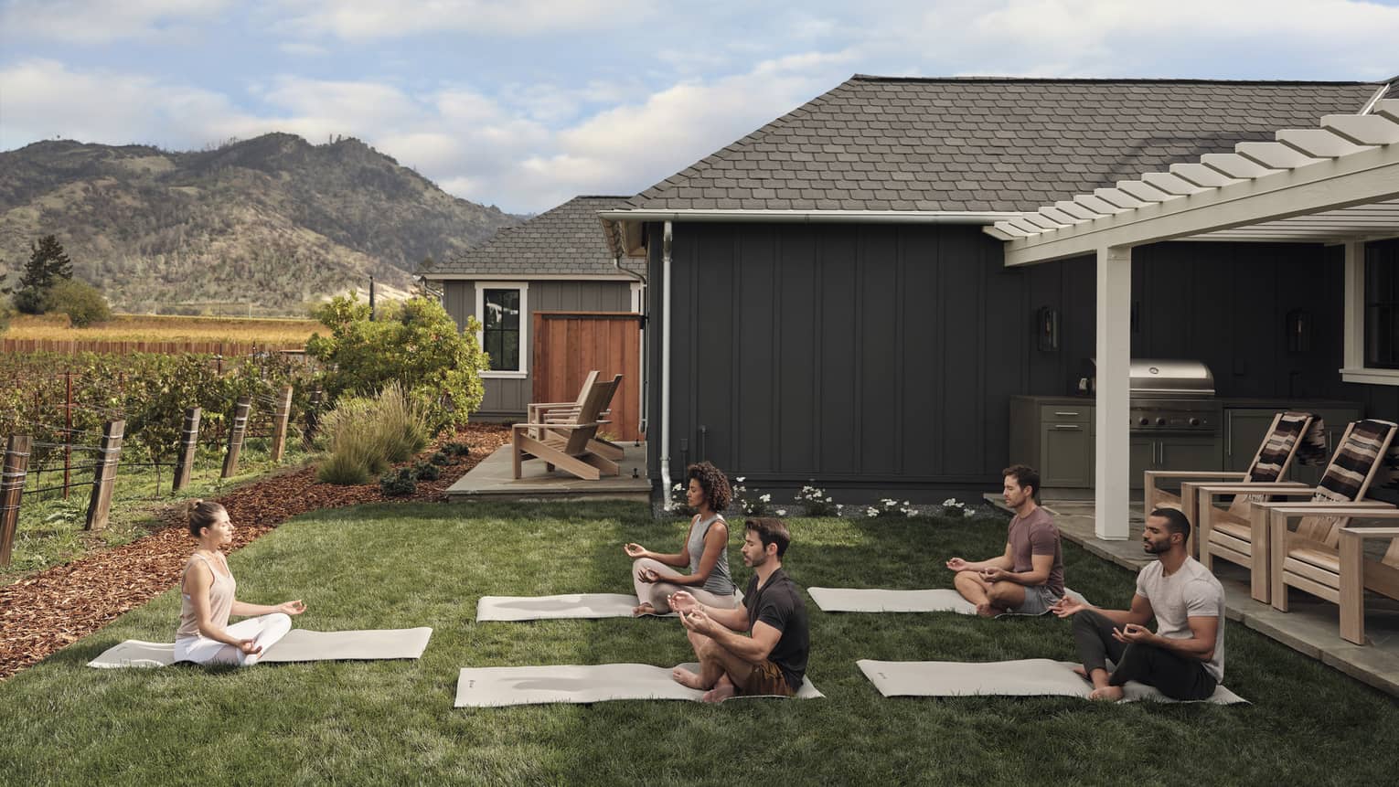 Group of people practicing yoga on mats in a grassy villa backyard near vineyards, with mountain views, wooden chairs and an outdoor grill area under a pergola