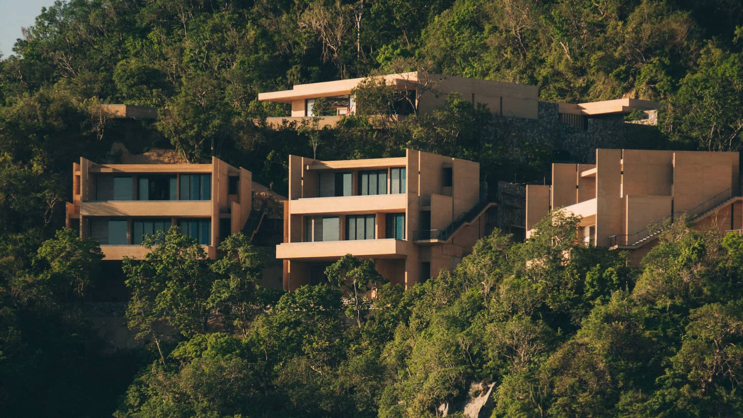 Modern cliffside villas surrounded by lush greenery, overlooking a secluded beach and ocean. The homes feature clean, angular designs, blending into the natural landscape.