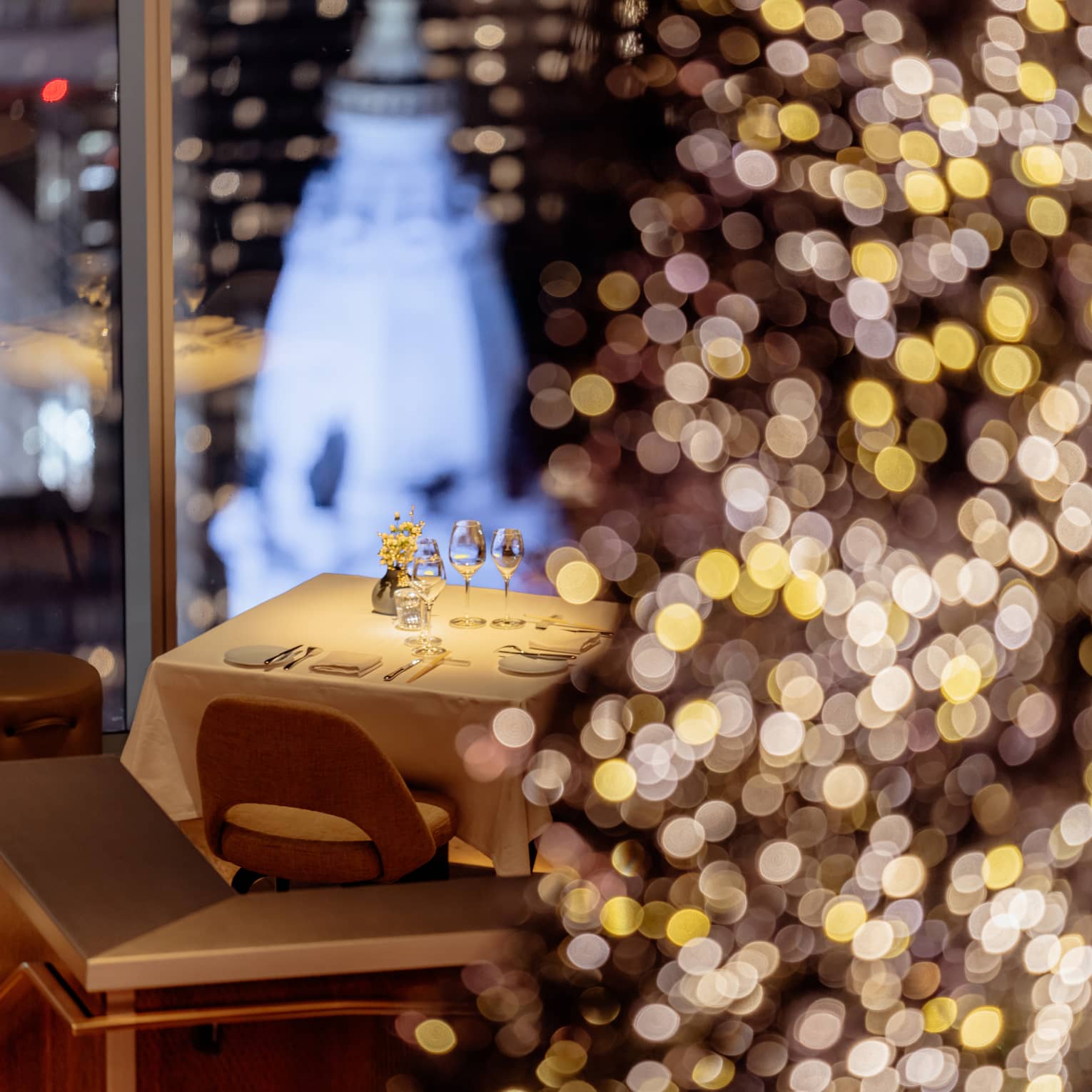 Restaurant table set for two next to floor-to-cieling windows overlooking a city skyline with a Christmas tree twinkling in the foreground