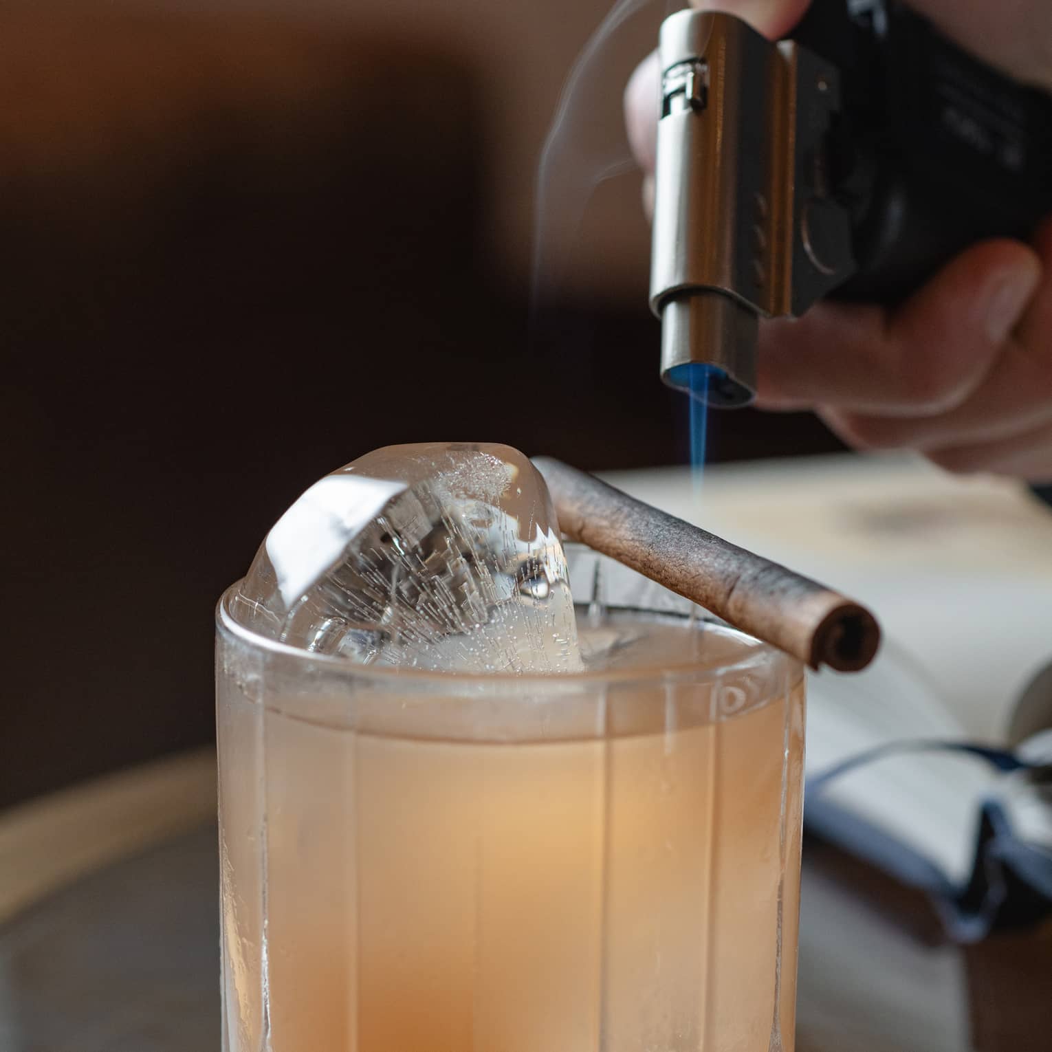 A hand holds a small blowtorch over a cinnamon stick set across the rim of a bowler glass of light amber liquid on ice.