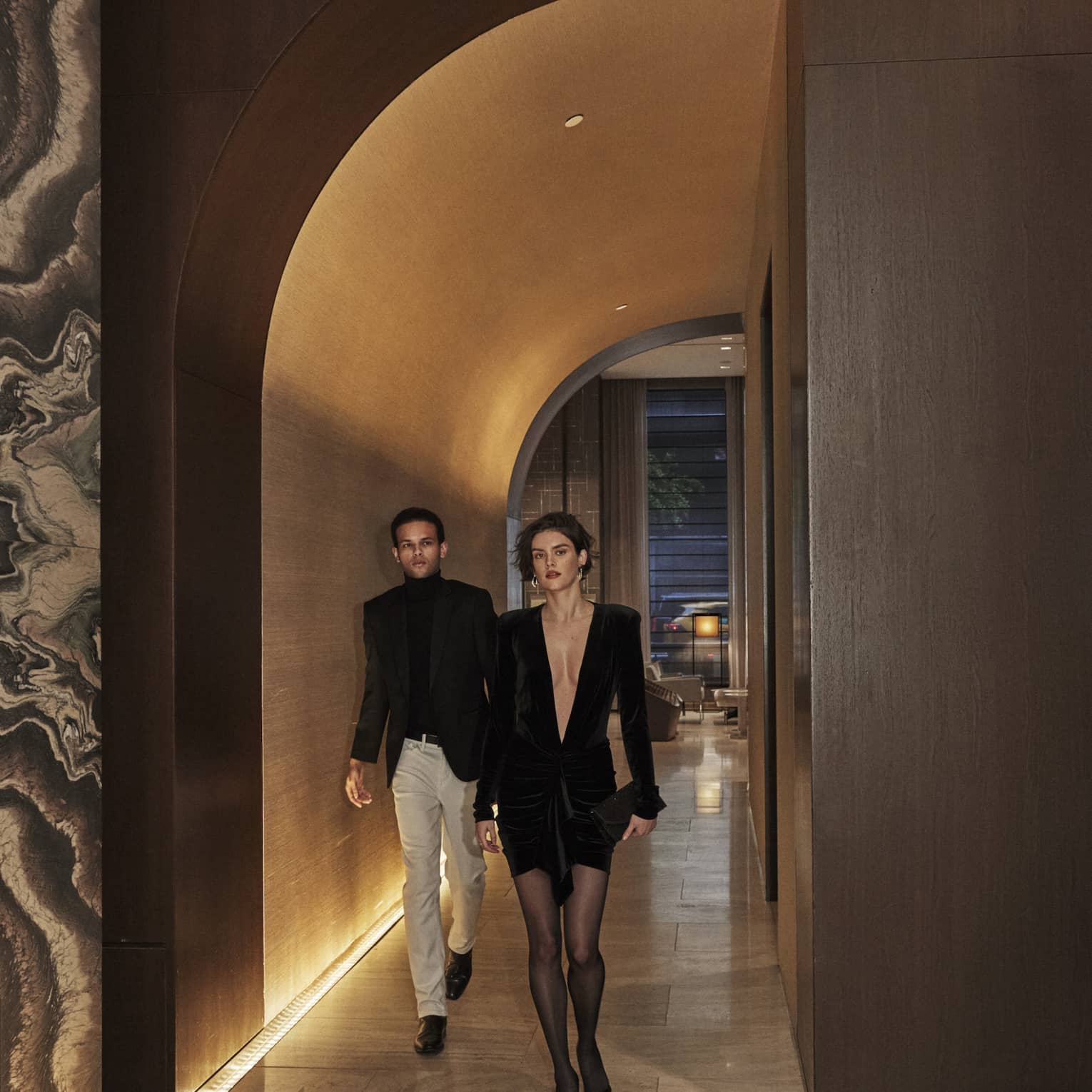 Elegantly dressed couple walking through a dimly lit, luxury hotel hallway with arched ceiling and marble accents