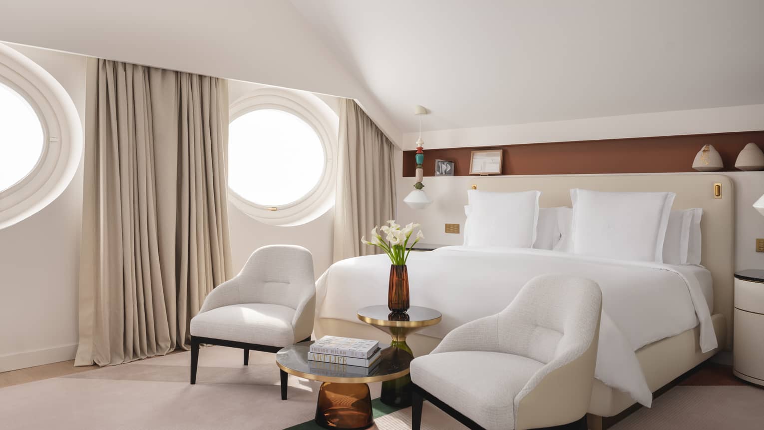 Contemporary bedroom with king bed, two ivory-coloured chairs at the foot of the bed, and two circular windows