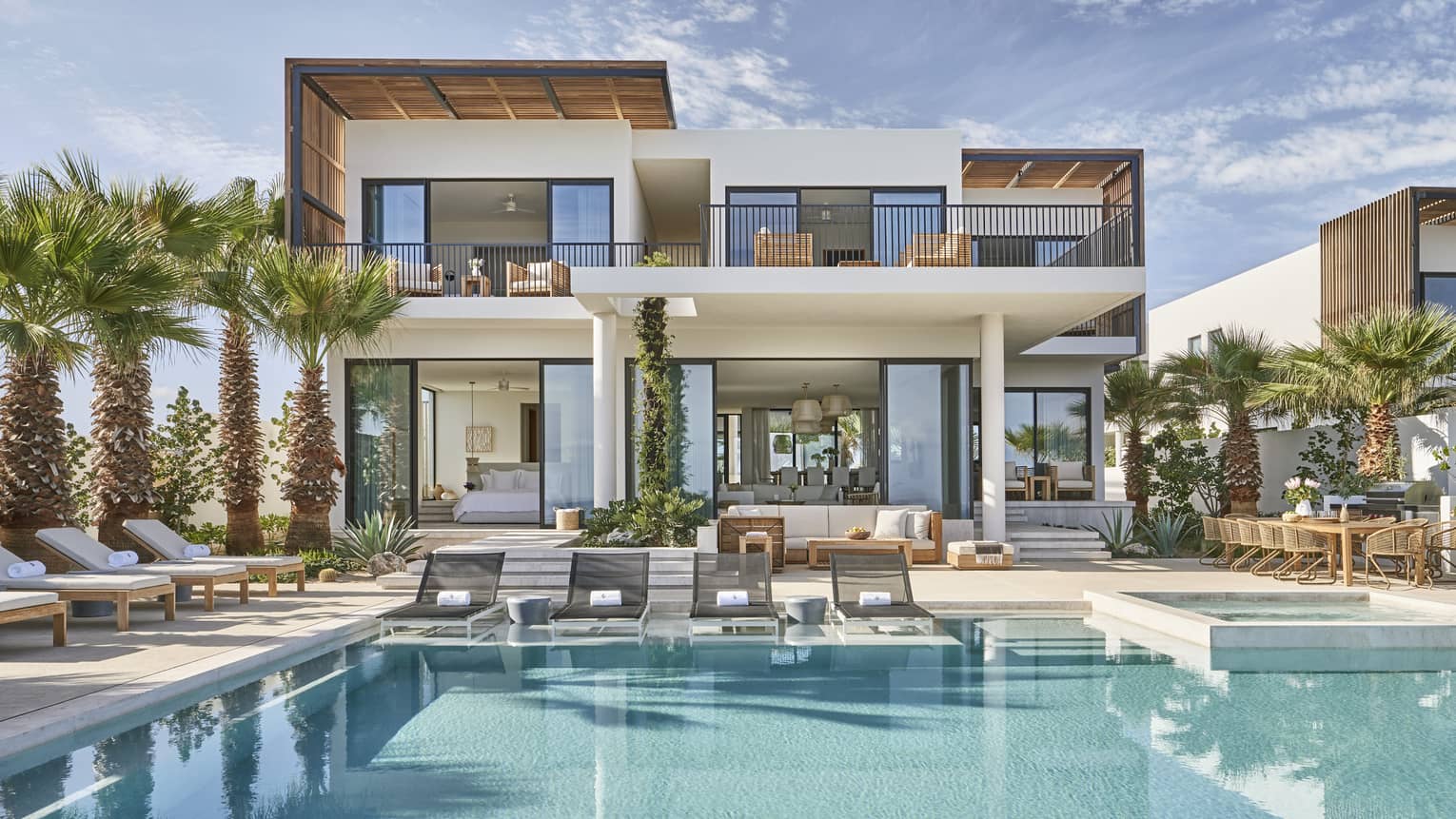 Modern luxury villa with a spacious pool area, surrounded by palm trees, featuring outdoor lounge chairs, a dining area, and floor-to-ceiling windows.
