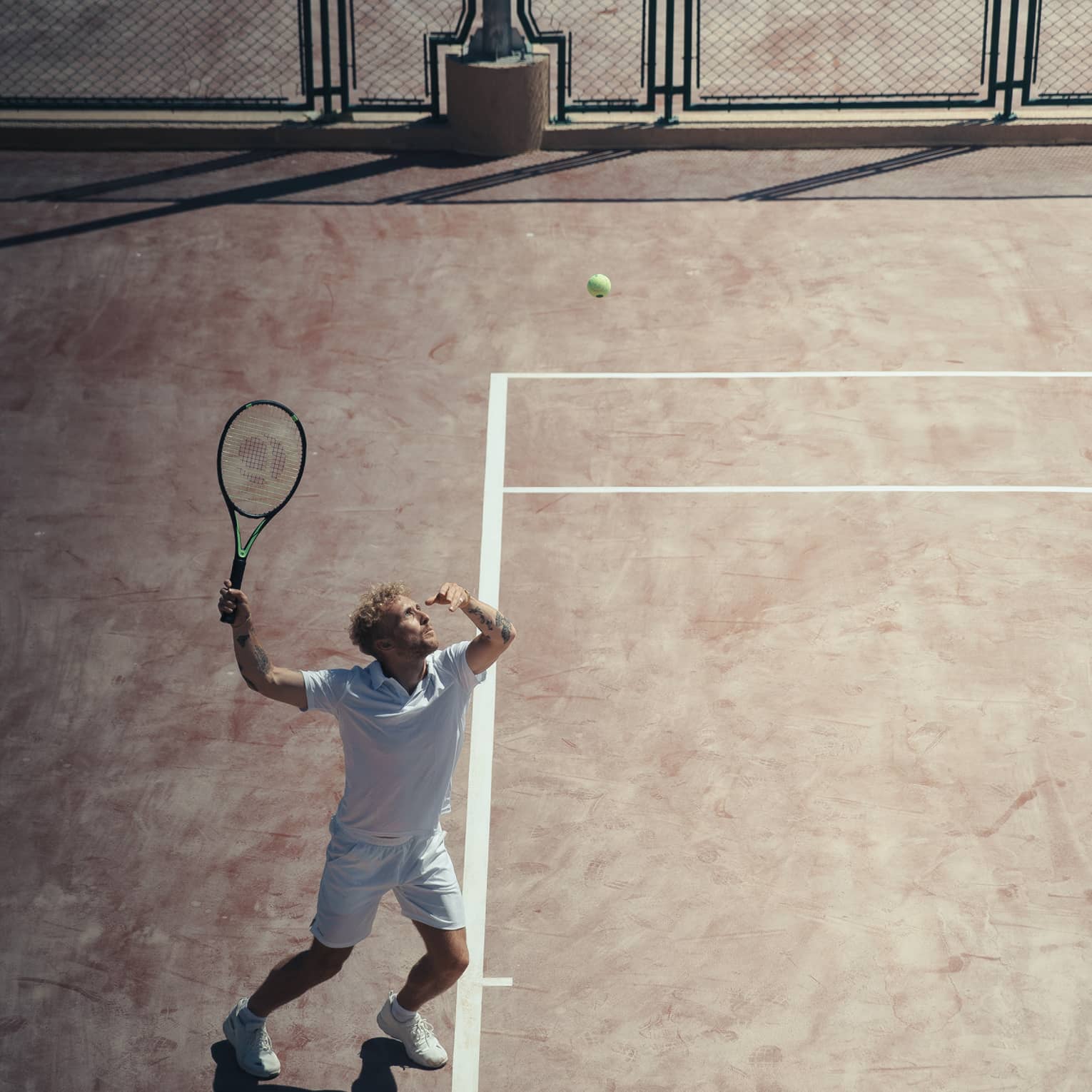 Man in white outfit playing tennis on reddish clay court