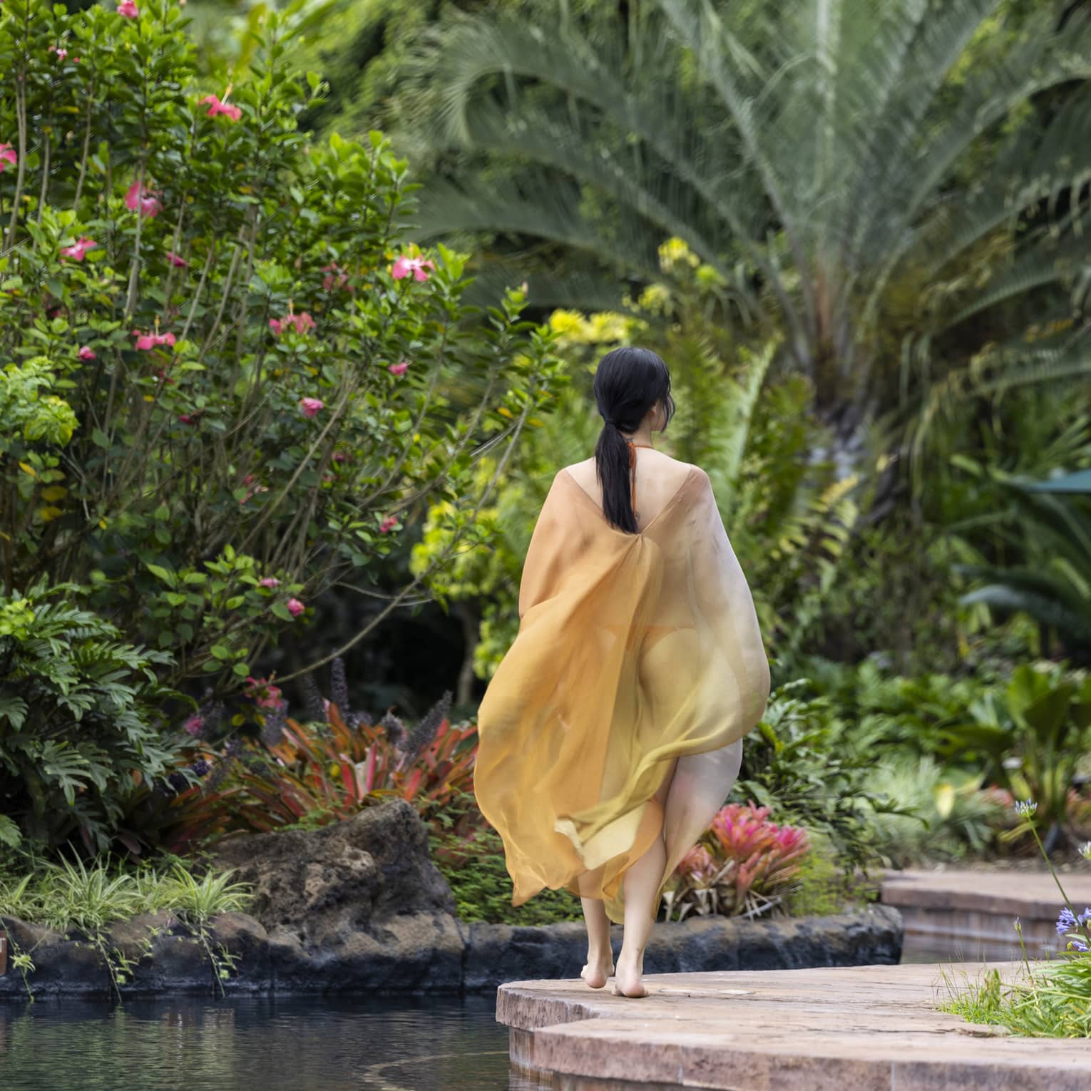 Rear view of a guest wearing a billowing silk cover-up walking along a path bordered by water, lush foliage and pink flowers.