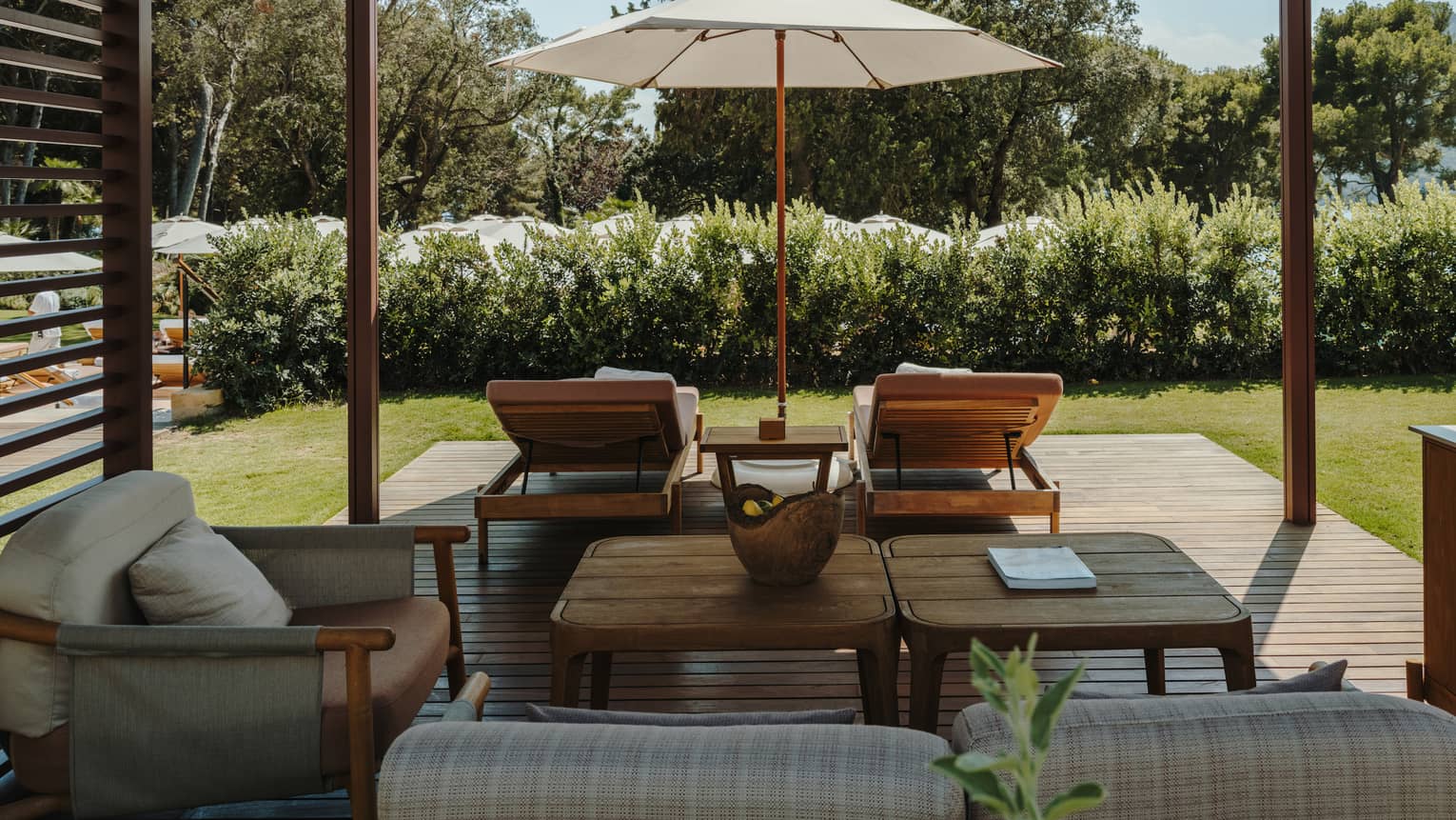 A shaded pool cabana with comfortable seating, a table, lounge chairs, an umbrella and lush green surroundings.