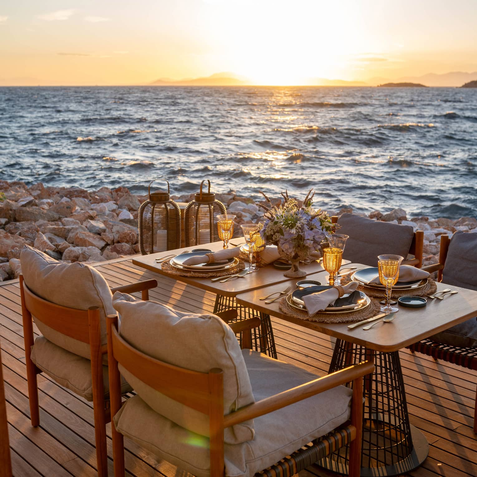 A table laid with sparkling glasses and a bouquet of flowers set on a deck on the rocky shore, the sunlit waves metres away.
