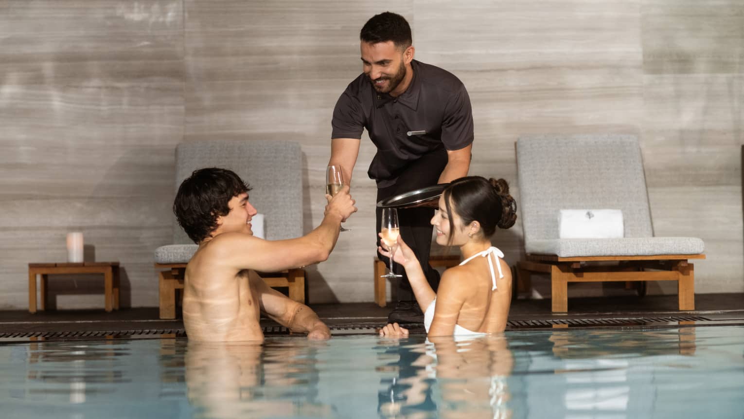 Two smiling guests in an indoor pool with glassy water and grey marble walls are served drinks in champagne flutes.