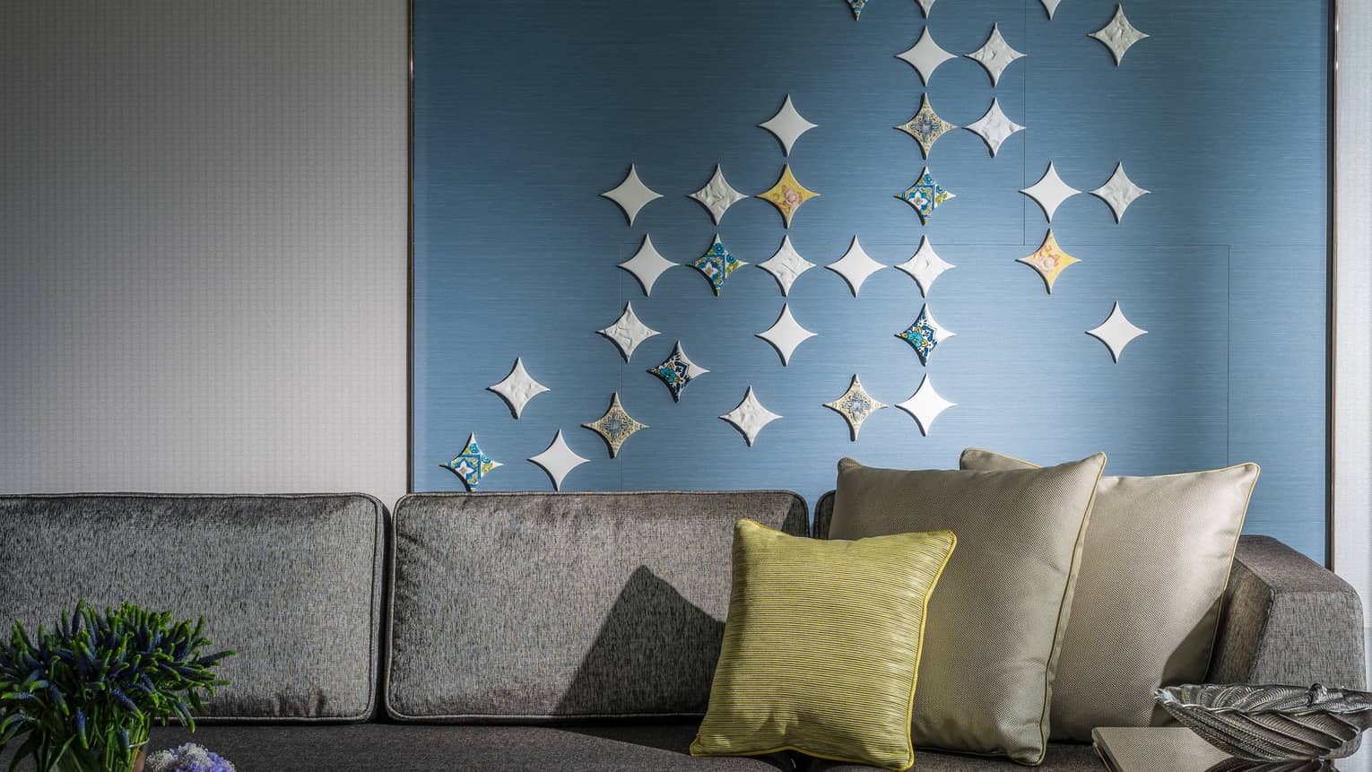 A modern living area featuring a grey sofa with decorative pillows, a small table holding flowers and books and a blue wall adorned with star-shaped accents.