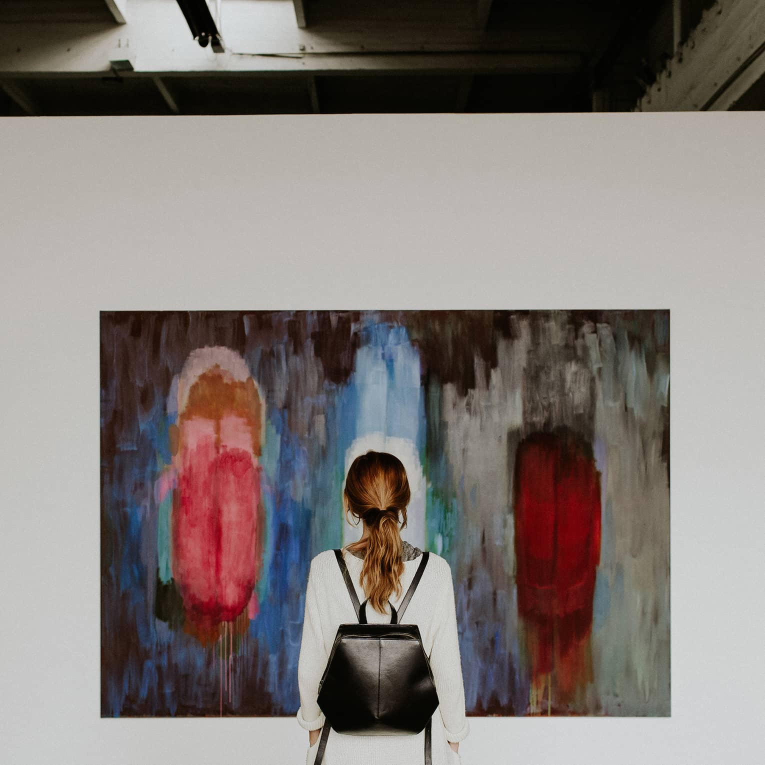A person with a backpack standing in front of an abstract painting in an art gallery.