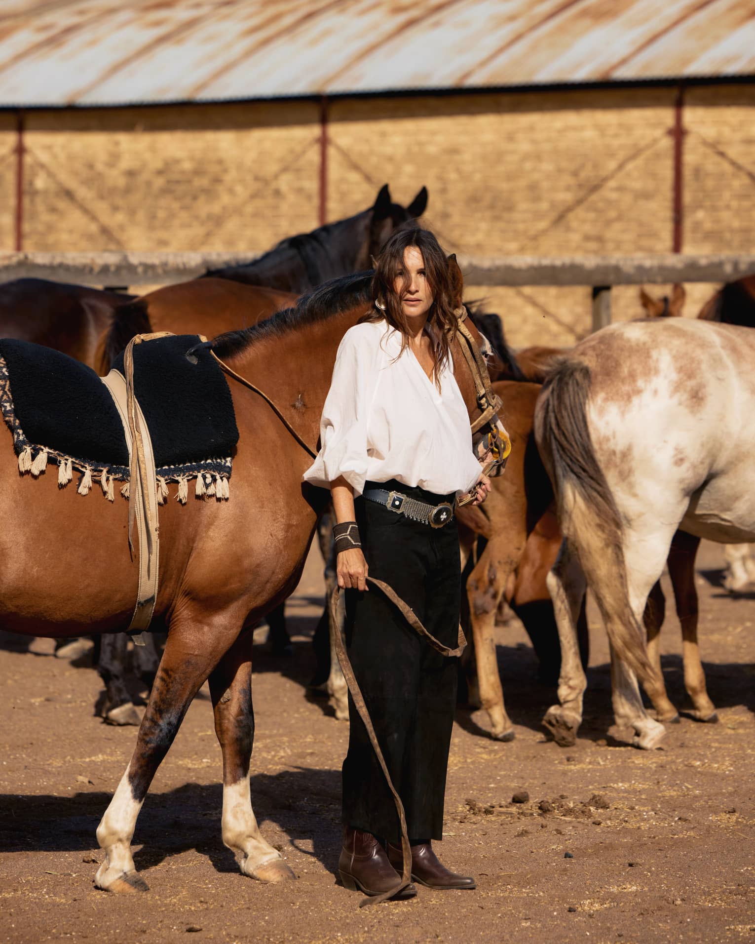 Woman with long dark hair wearing a loose-fitting white button-down shirt, black pants, brown boots and a large belt stands holding the reins of a brown horse with multiple other horses in the background