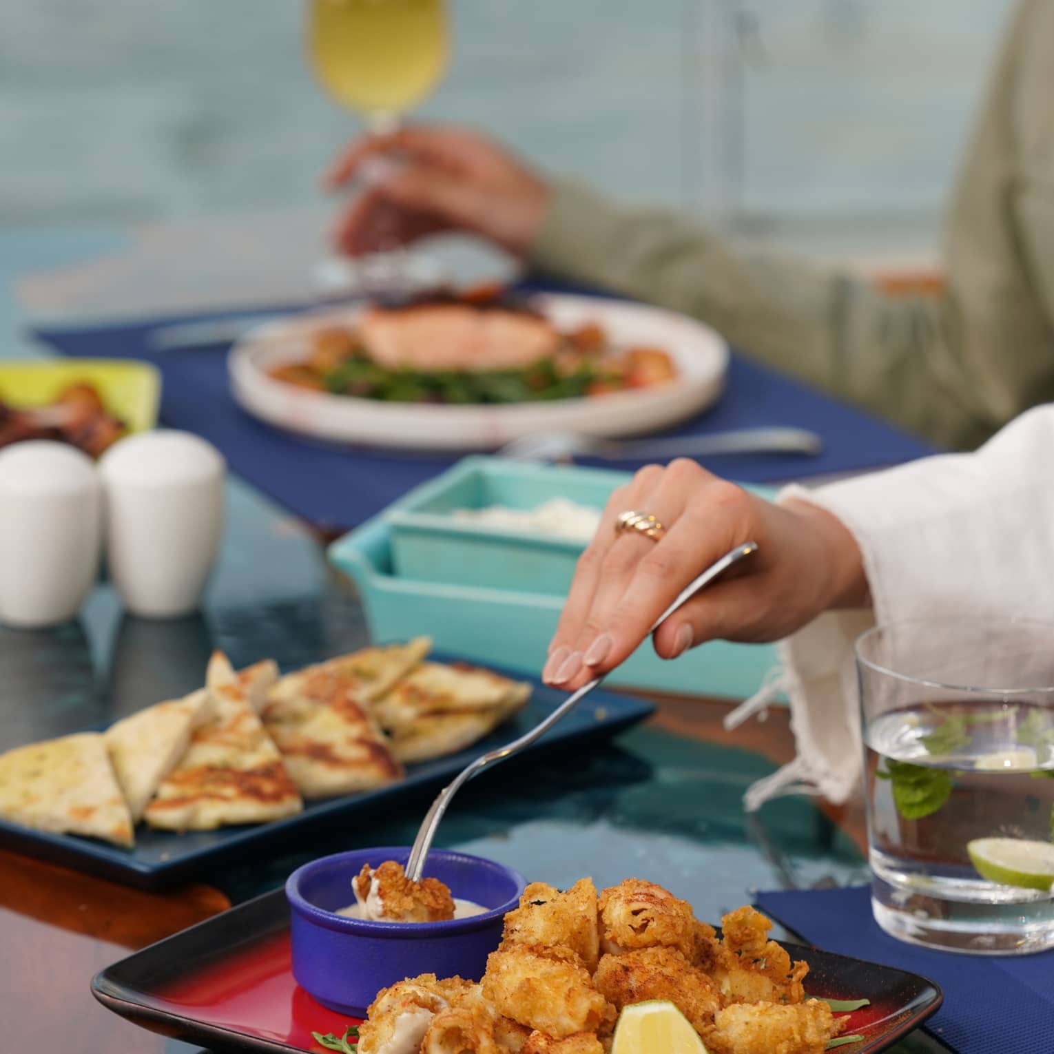 Hands dips morsel of food into small blue bowl of sauce on platter of fried seafood