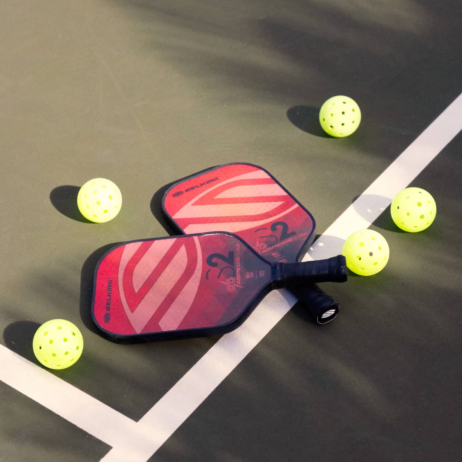 Close-up of two red and pink pickleball racquets, handles crisscrossed on a white court line, surrounded by five pickleballs.