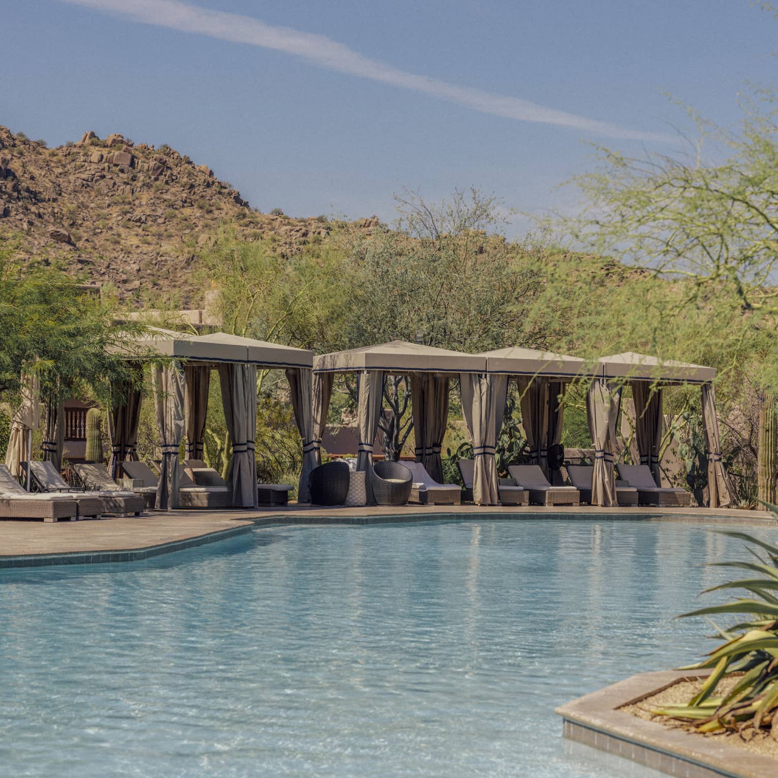 Four cabanas with lounge chairs near the pool with mountains in the background.