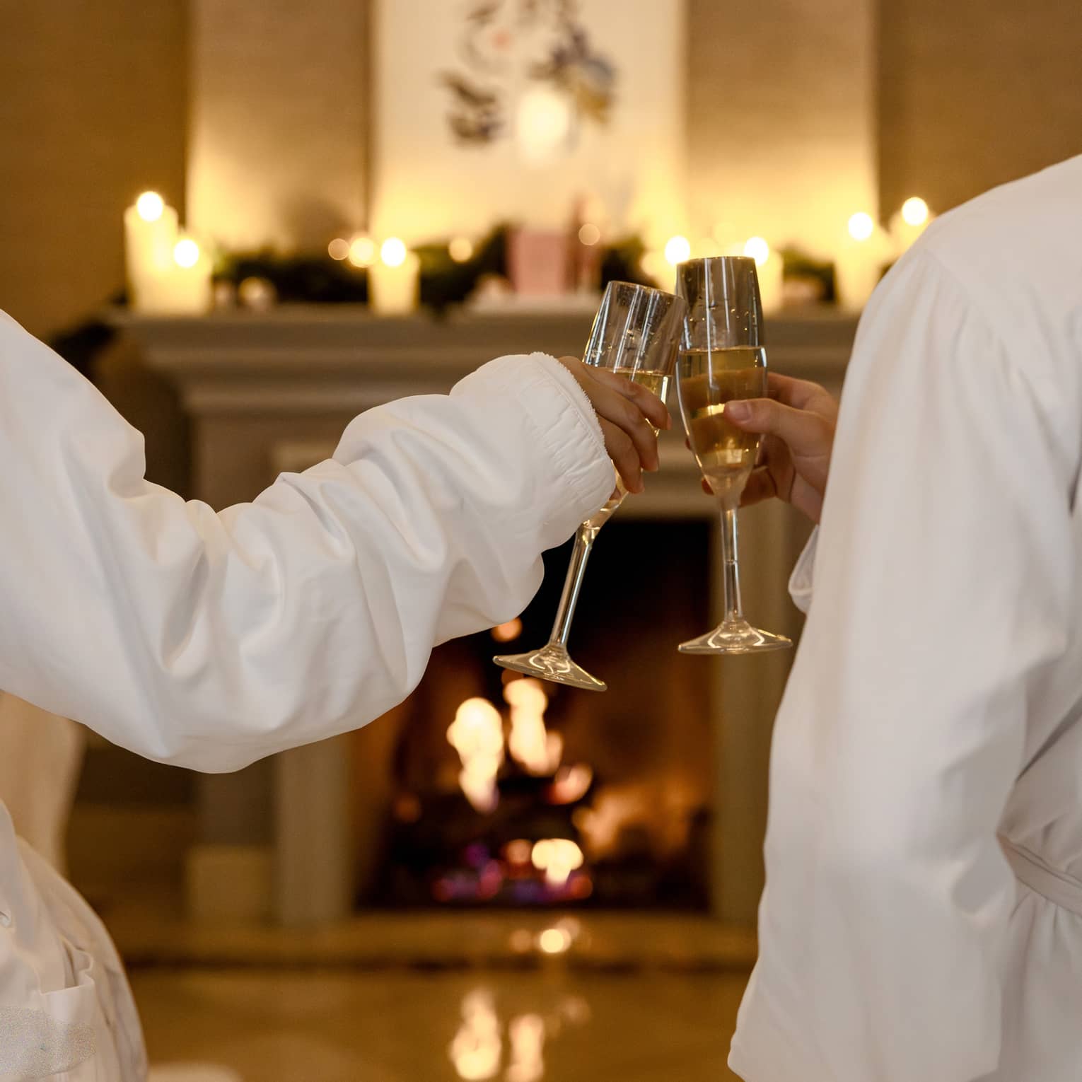 Two guests wearing white robes sit in front of a fireplace toasting each other with glasses of Champagne