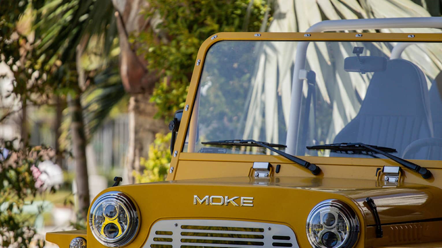 Extreme close-up of a dark-yellow Moke parked in the shade against a backdrop of palm trees and other greenery.