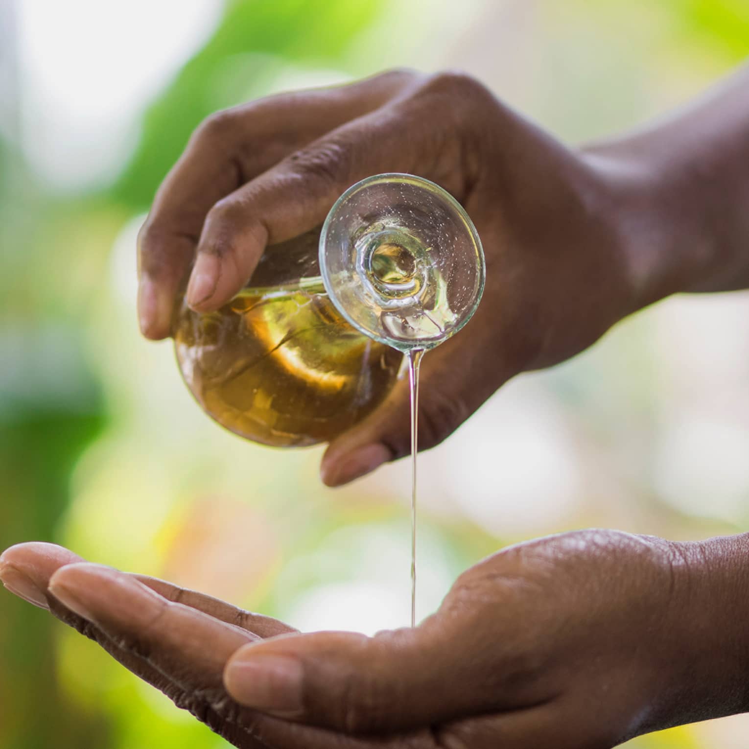 Golden oil flows from a small carafe held in one hand onto the other, a small wooden bowl underneath to catch the overflow.
