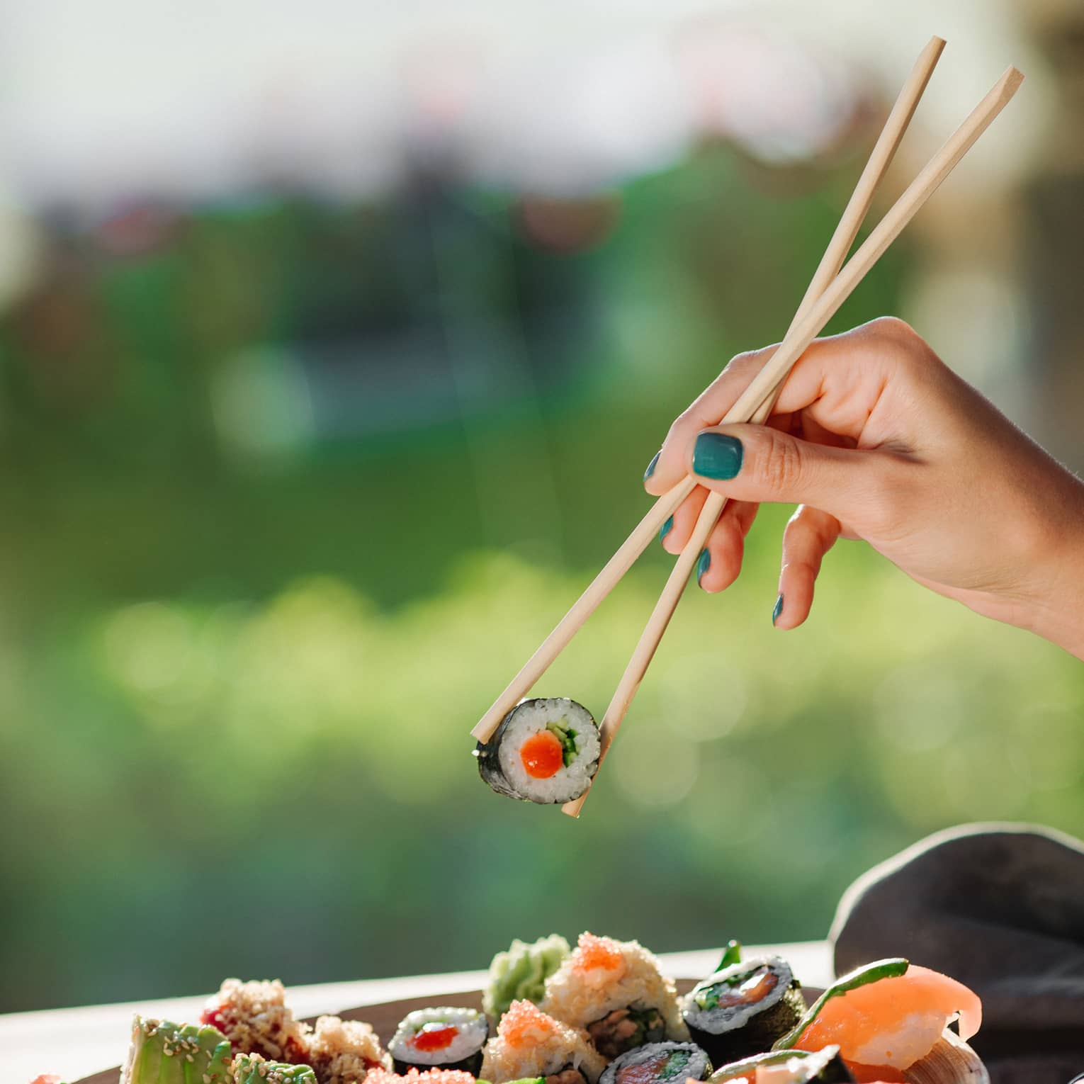 Hand pinching chopsticks, holding a piece of sushi over a plate of assorted sushi