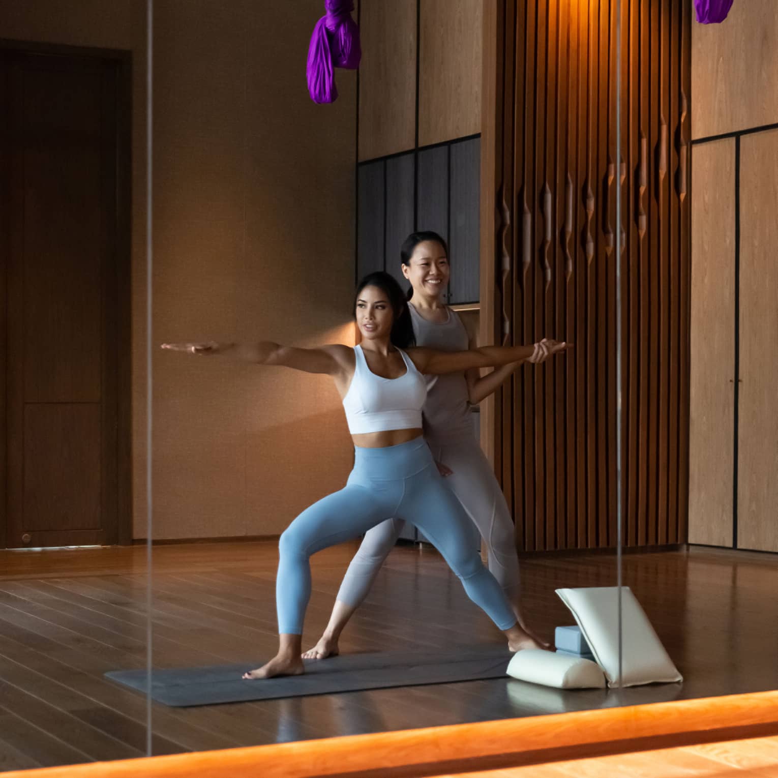 Mirror reflection of fitness staff helping woman stretch her arms in yoga pose under purple ribbons