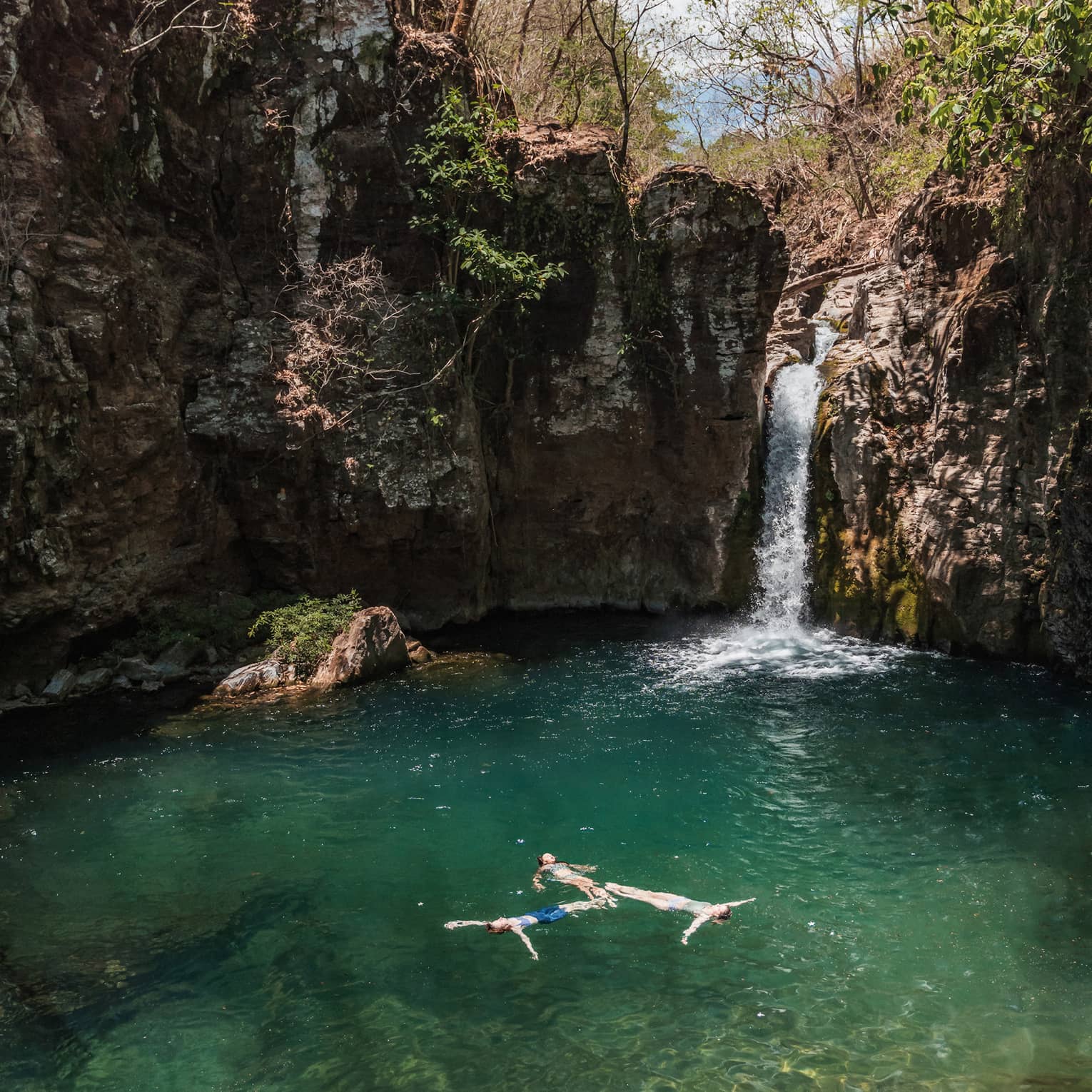 Three people float in a lagoon near a waterfall