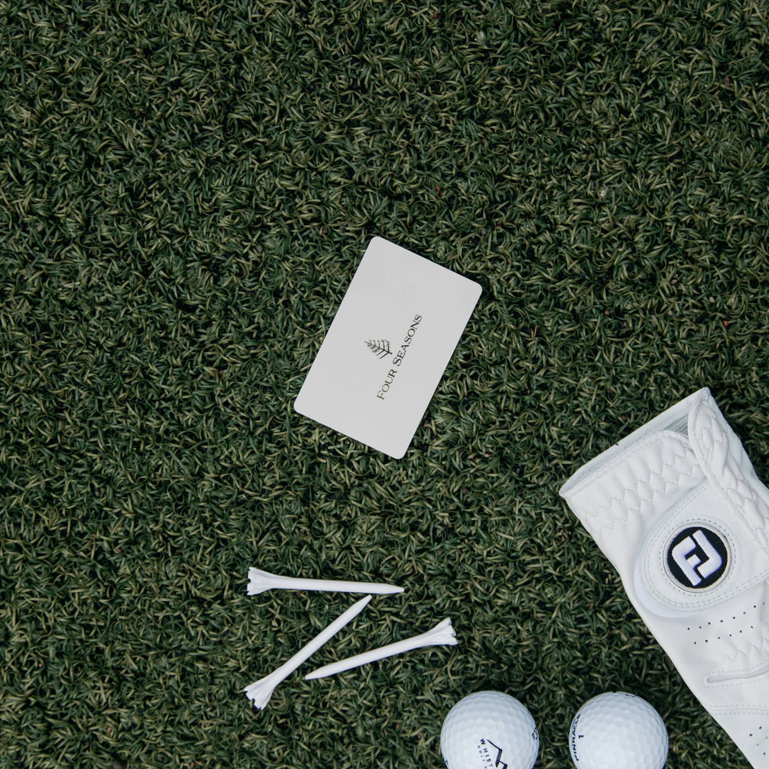 On green turf, a Four Seasons card, three golf balls, three tees and a white leather golf glove dotted with tiny holes.