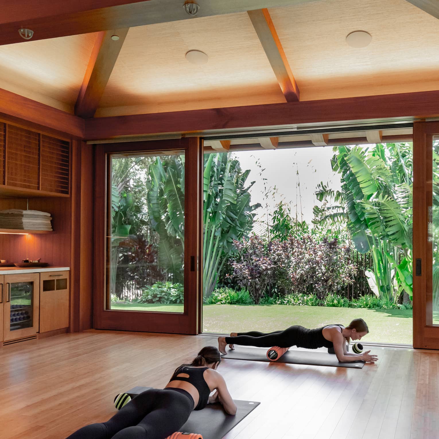 A sunny studio with exposed wooden beams opens to a lush garden. Inside, two fitness enthusiasts foam roll on exercise mats.