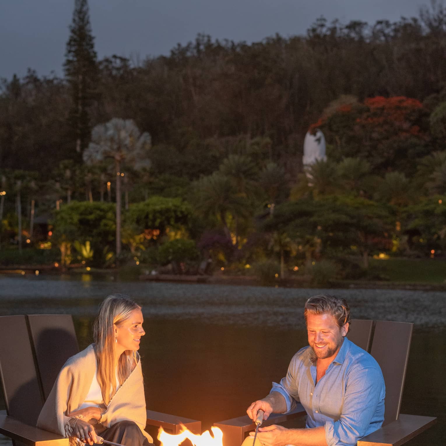 Couple in large lounge chairs beside a lake roast marshmallows over a round propane firepit table with black fireglass.