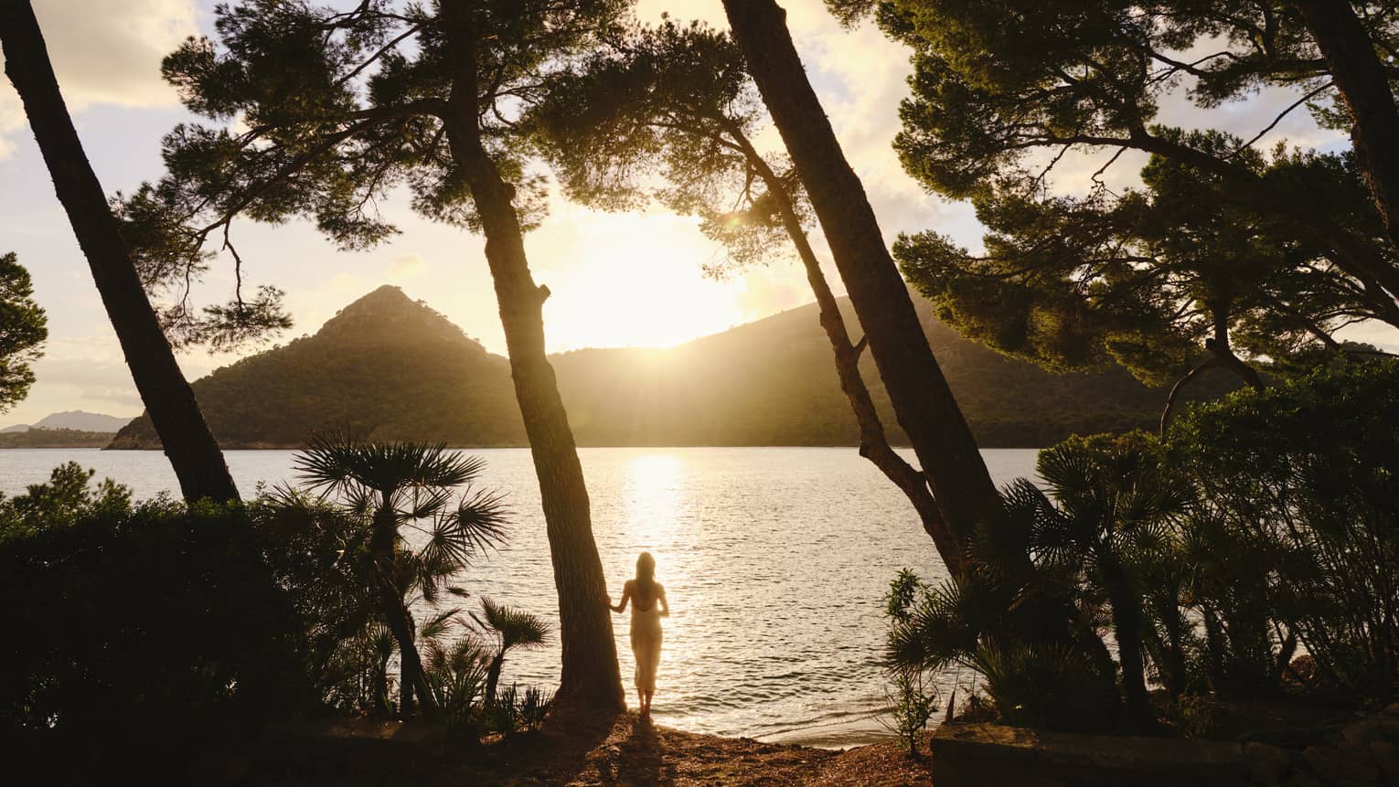 Silhouetted by the sun dipping behind a mountain, a guest stands at the edge of rippling water framed by tall leaning trees.