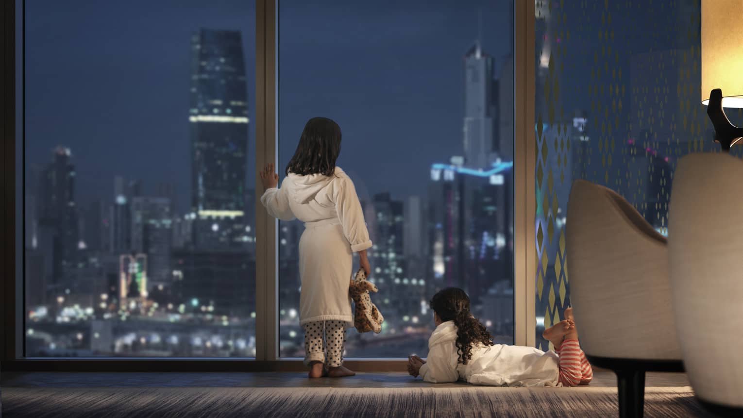 Two young children wearing bath robes look out a floor-to-ceiling window at a nighttime view of an urban cityscape.