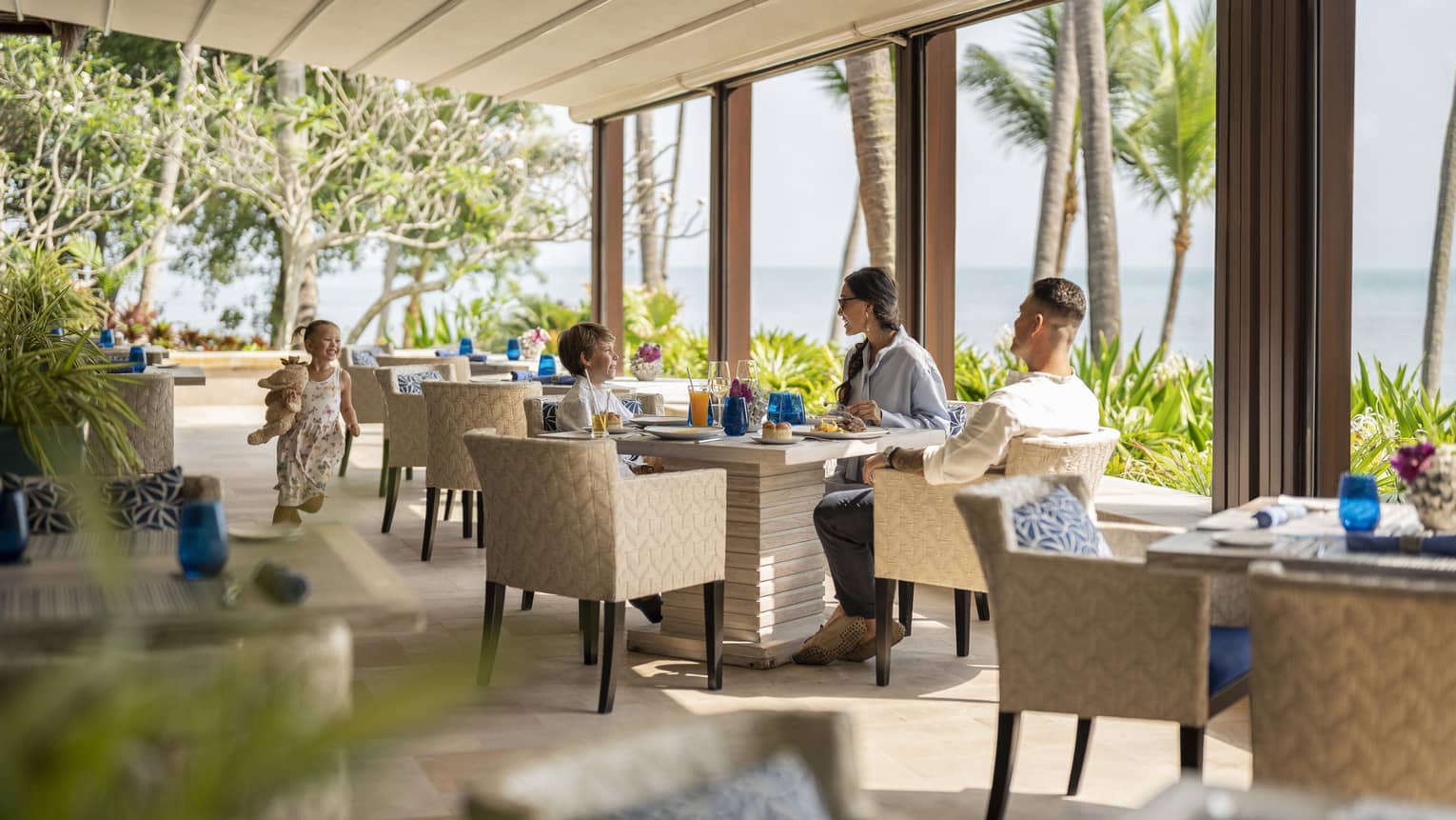 Two parents and a child eat under a covered patio next to the ocean. Their youngest child runs toward them from the left.