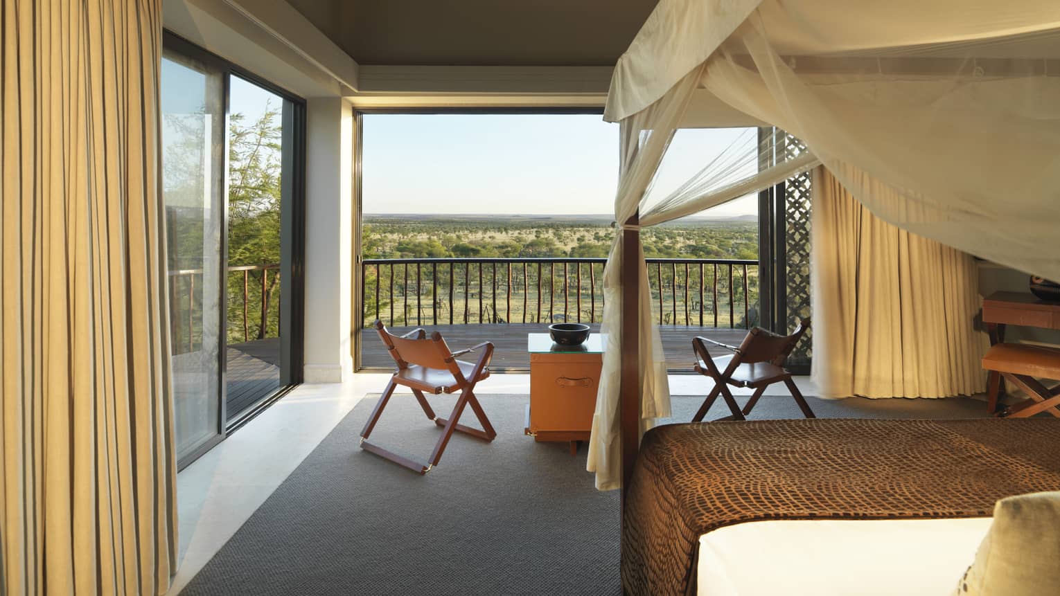 Poster bed, canopy by fold-up chairs, wood table by open wall with Serengeti views