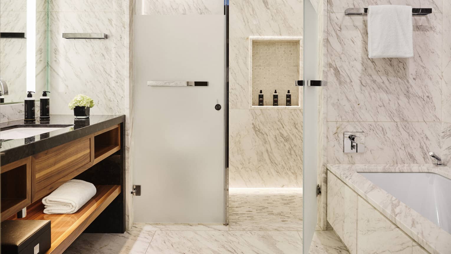 Luxury marble bathroom with walk-in shower, bathtub, vanity with black countertop and wood shelving, towel rack and toiletries displayed on a recessed shelf