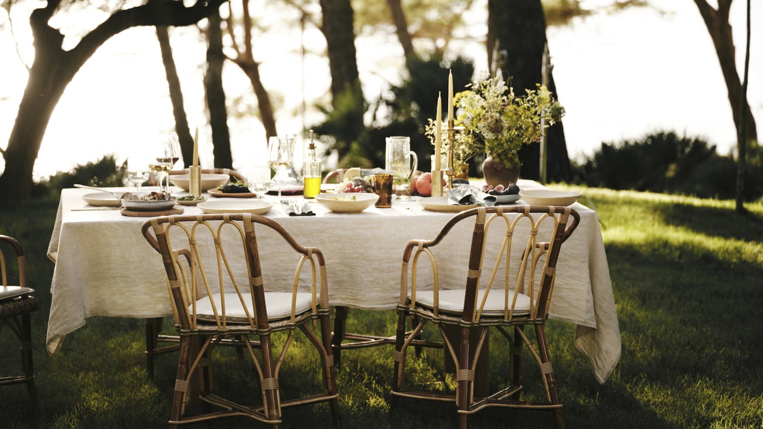 A linen tablecloth adorns an outdoor table set for five with wine, fresh seafood and fruit in an ocean-view forest setting.