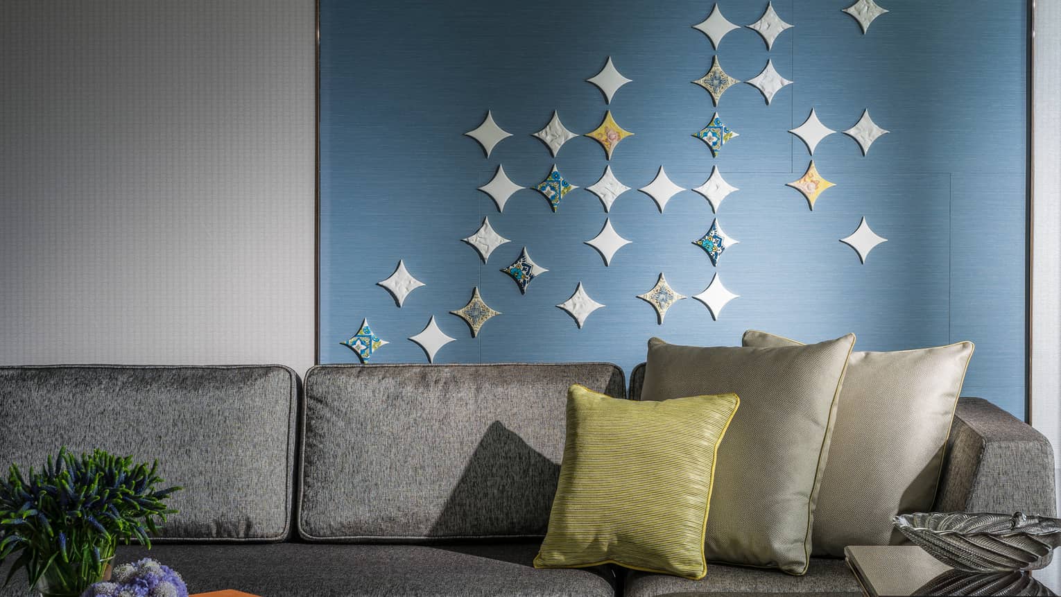 A modern living area featuring a grey sofa with decorative pillows, a small table holding flowers and books and a blue wall adorned with star-shaped accents.