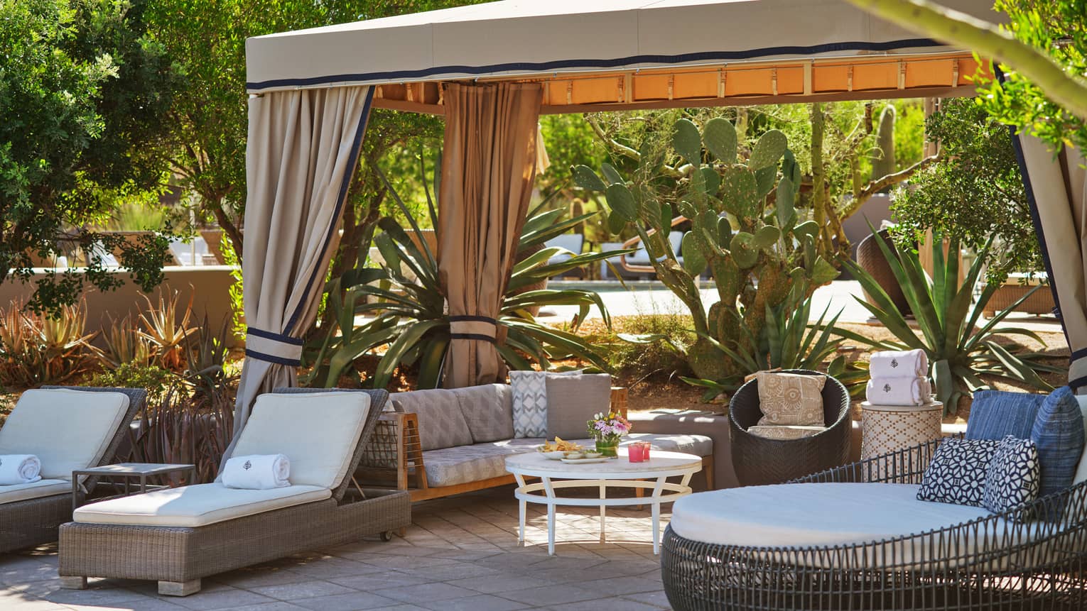 Cabana with trees and greenery surrounding, two brown and white lounge chairs and a couch.