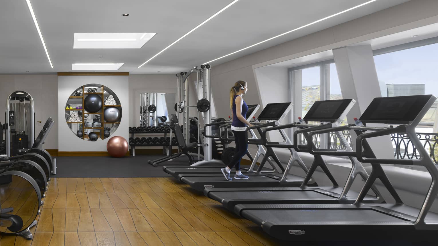 A guest walks on one of a bank of treadmills facing windows in a bright, modern gym with a striking geometric wood floor.