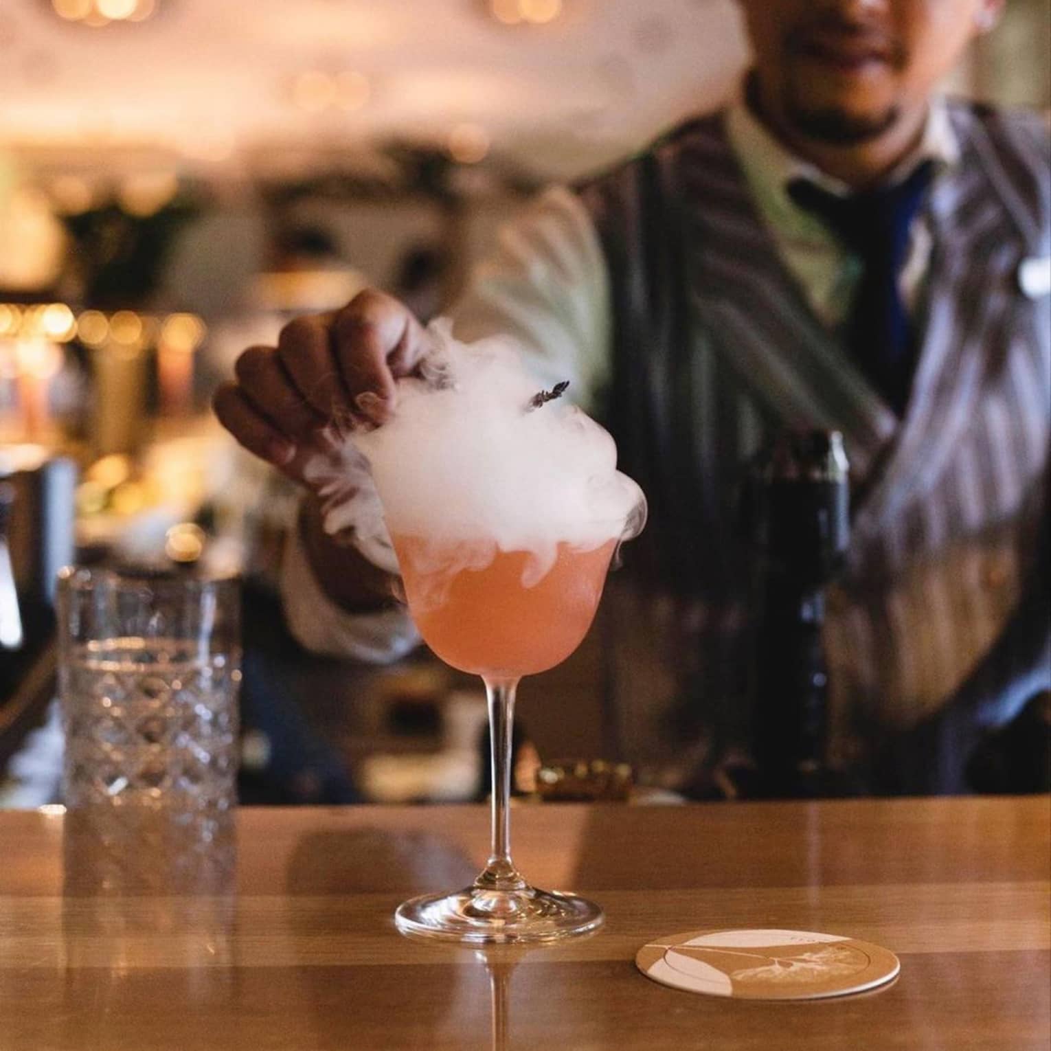 A mixologist garnishes a bright orange cocktail atop a smooth wooden bar; smoke billows over the sides of the stemmed glass.
