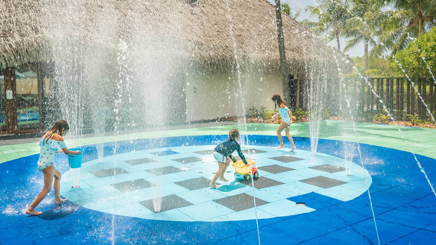 Group of children in swimsuits play on splash pad next to grass-roof building