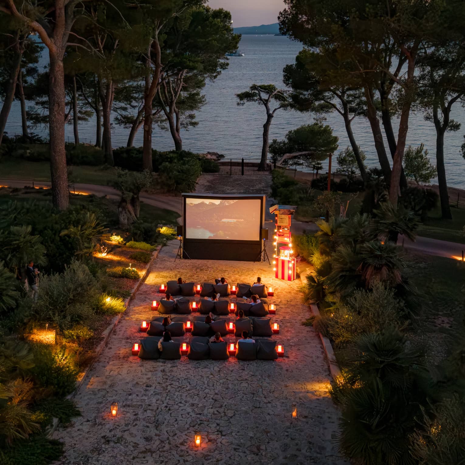 A bird’s-eye view of people seated in rows of chairs in front of a large movie screen in an outdoor setting in the evening, surrounded by trees, with the ocean in the background