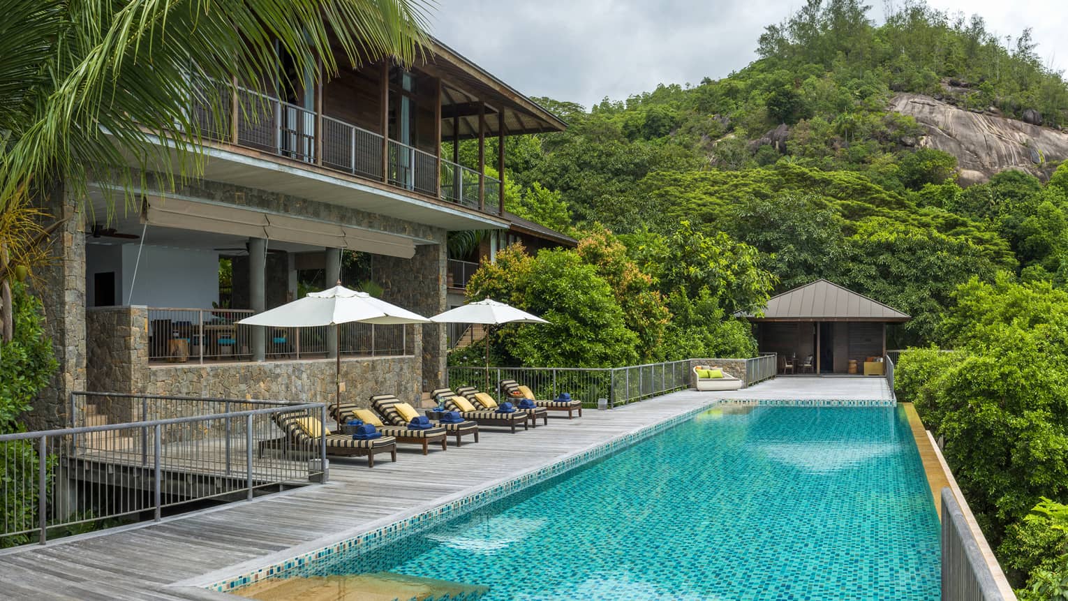 Row of patio chairs and umbrellas on balcony beside long blue swimming pool, mountain in background