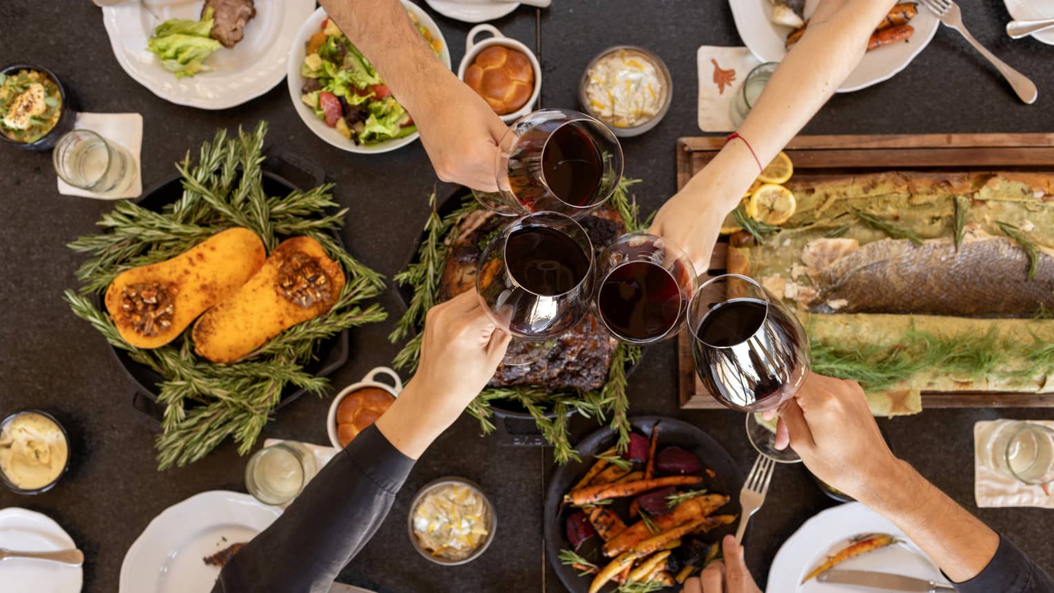 Overhead view of a table filled with festive dishes such as roasted squash, a filet of fish and other platters with four hands each holding a glass of red wine coming together in the centre