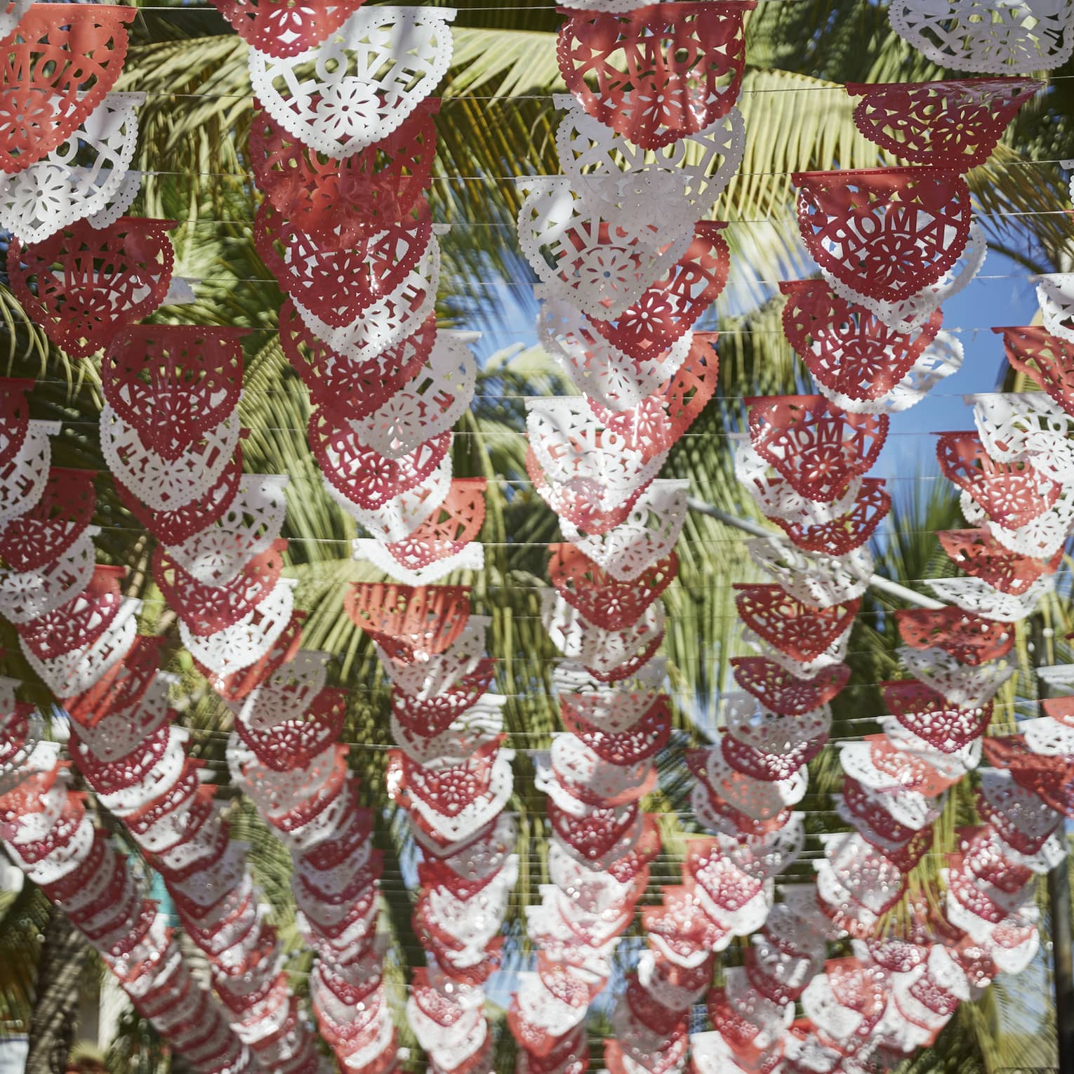 Looking up at rows of red and white Mexican Papel Picado banners hanging above, with palm trees and a sunny sky above them.