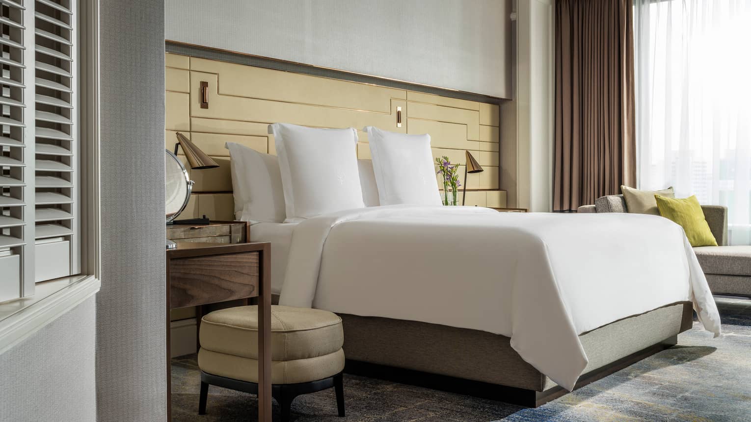 A bedroom with a large bed dressed in white linens, cushioned stool and wooden nightstand. Natural light streams through the window, brightening the space.