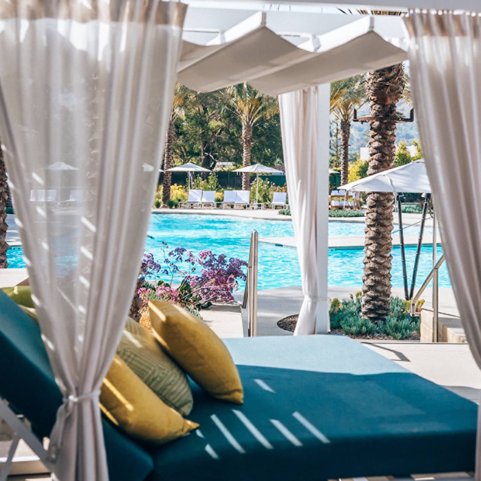 Poolside cabana featuring a blue daybed with yellow pillows and sheer beige curtains