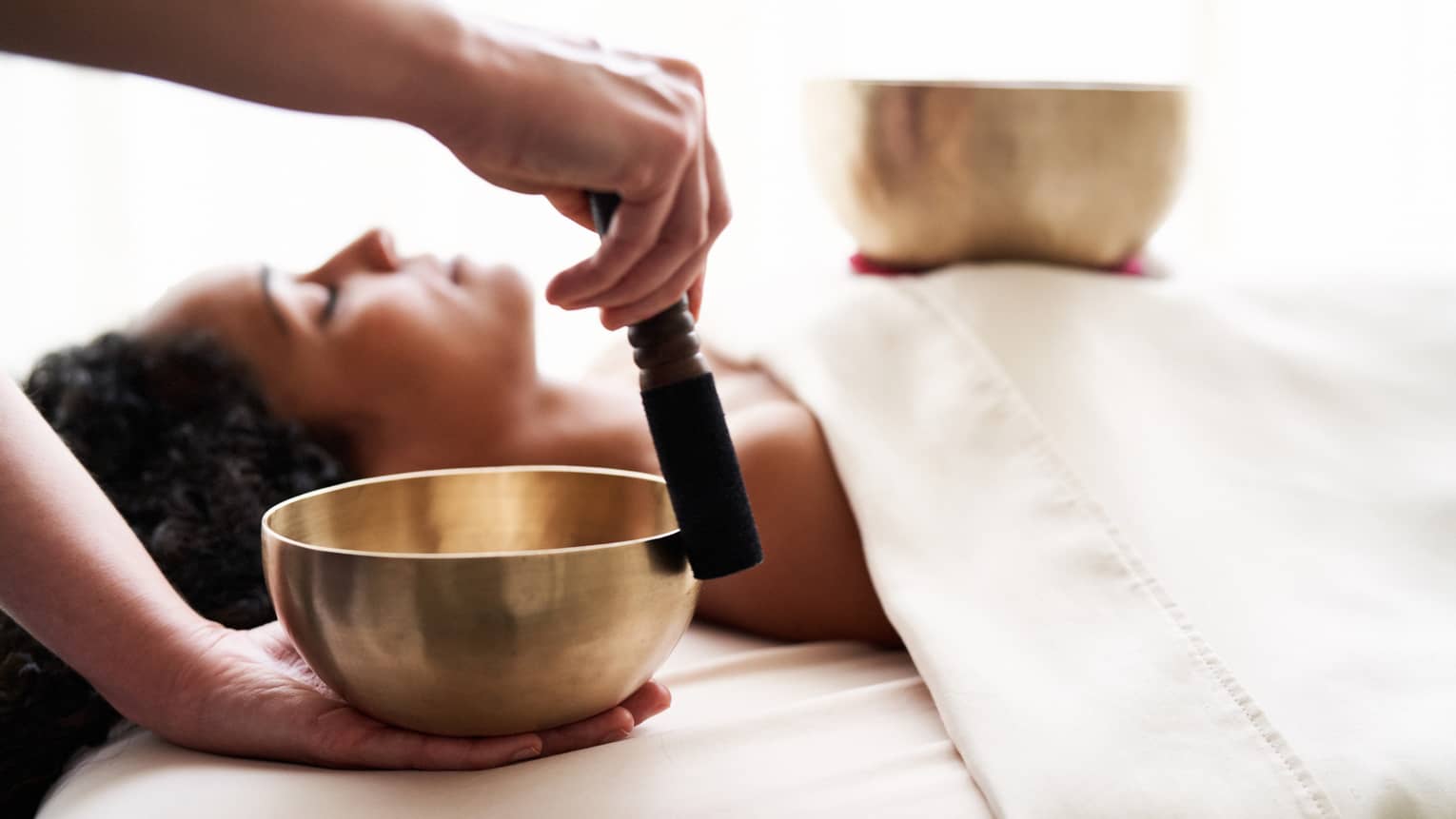 Woman with eyes closed lies on massage table in as vibrational sound therapy is performed with a Tibetan singing bowl