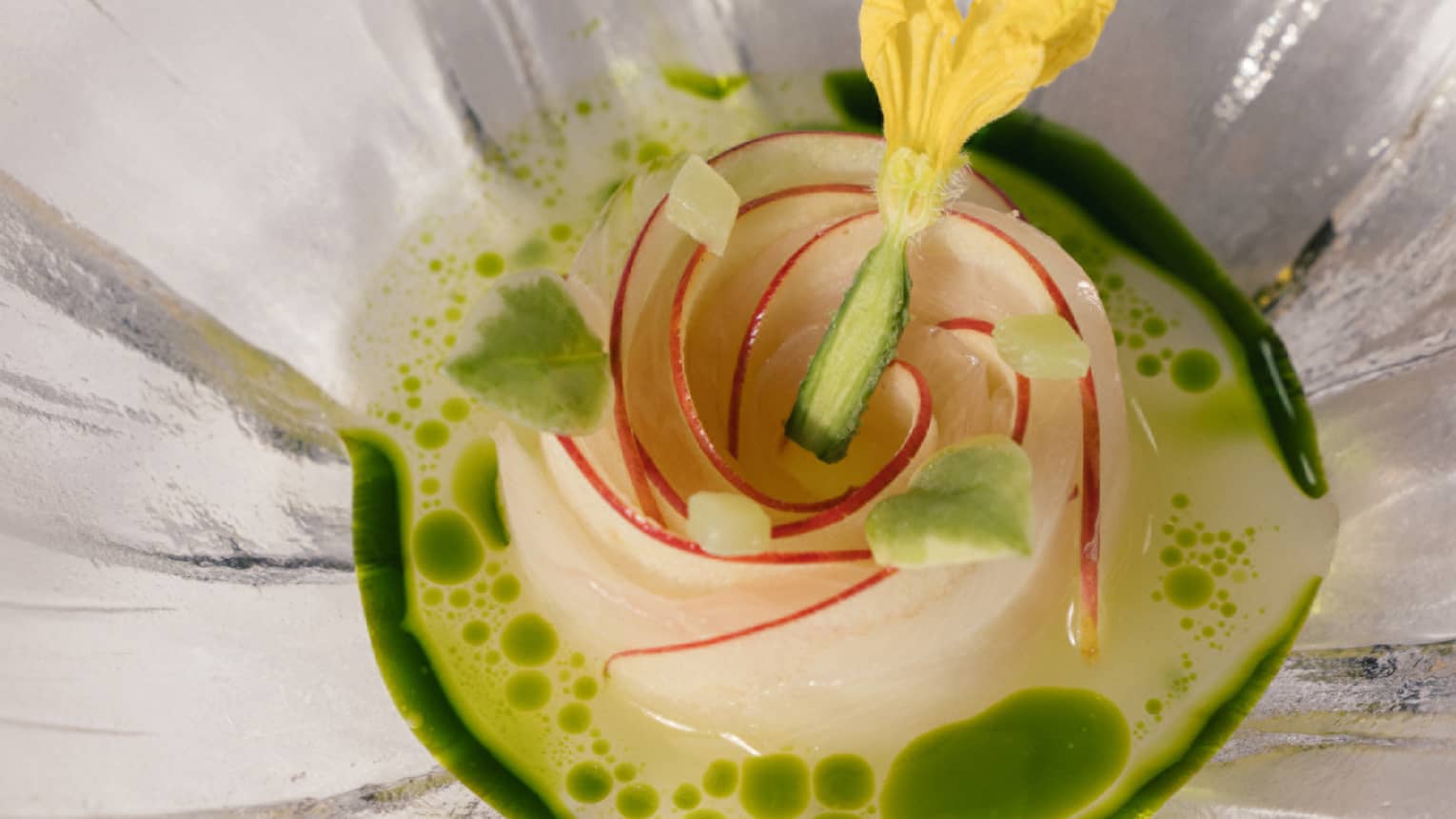 Elegant dish arranged as a rose, topped with a yellow edible flower, in a green herb-infused broth