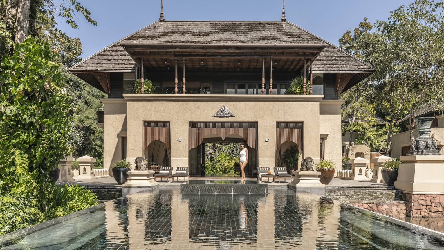 Person walking along the edge of a large reflective pool in front of a grand villa with traditional architecture surrounded by trees and outdoor seating