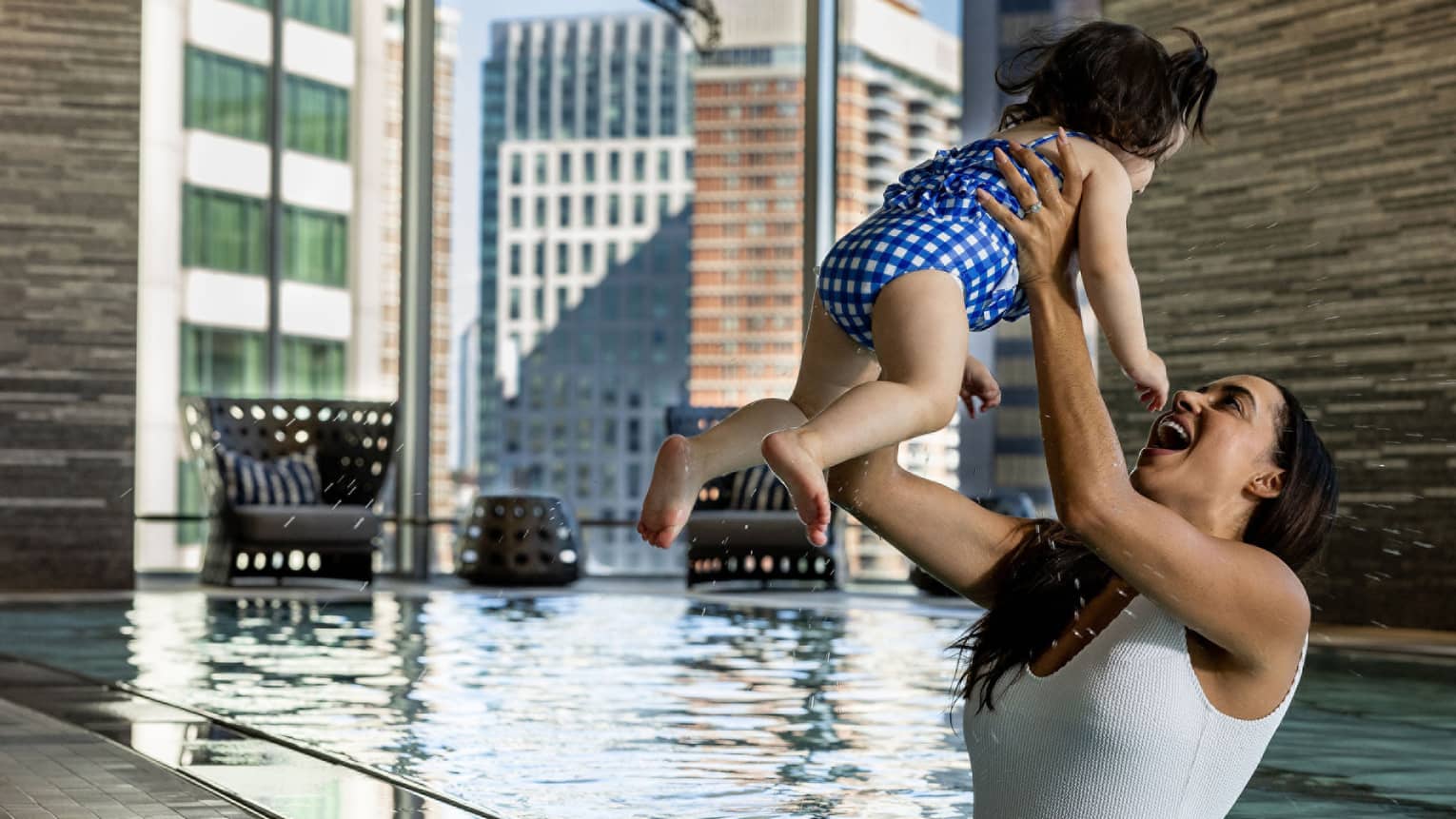 A smiling mother holds her toddler aloft in an indoor pool with floor-to-ceiling windows looking out onto the city.